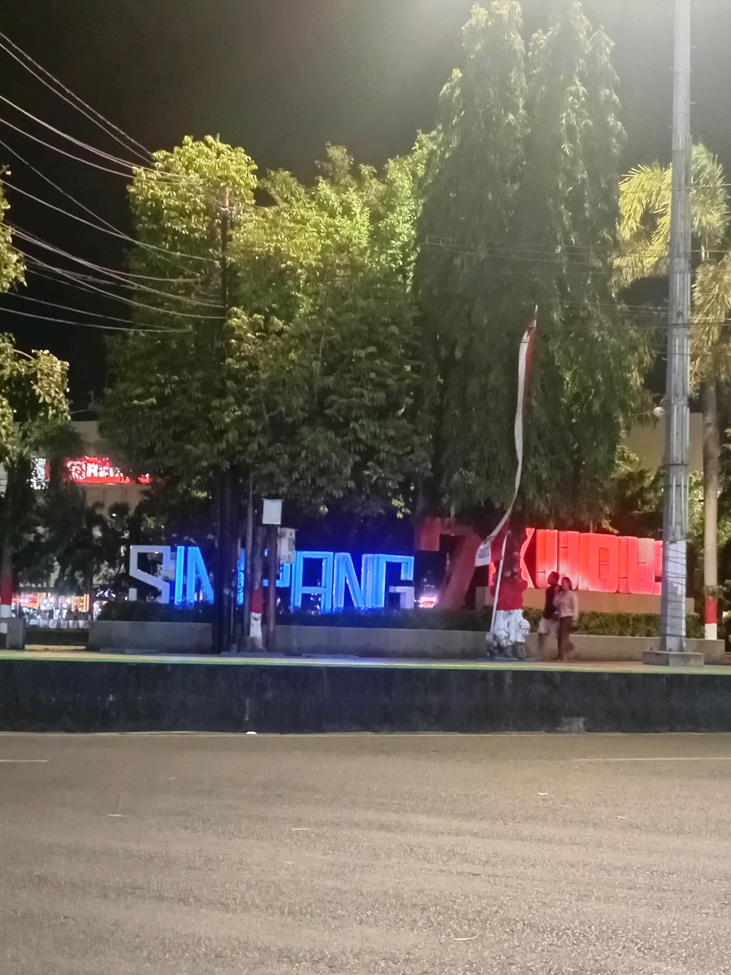 The image depicts a nighttime street scene in Kudus, Indonesia. Dominating the foreground is an asphalt road. Behind it, a raised area features large, illuminated blue letters spelling out "SIMPANG" on the left, and red illuminated letters on the right, partially obscured by trees and a flagpole with a red and white Indonesian flag. Several trees, some tall and slender with dense foliage, others shorter and more bushy, are scattered across the scene. Electrical wires are visible crisscrossing the upper left portion of the image. A street light illuminates the area, casting a glow on the foliage. Two people are visible in the mid-ground, near the flagpole, walking away from the camera. The background includes more trees and what appears to be a building with bright lights. The overall atmosphere is urban and illuminated by artificial lights.
