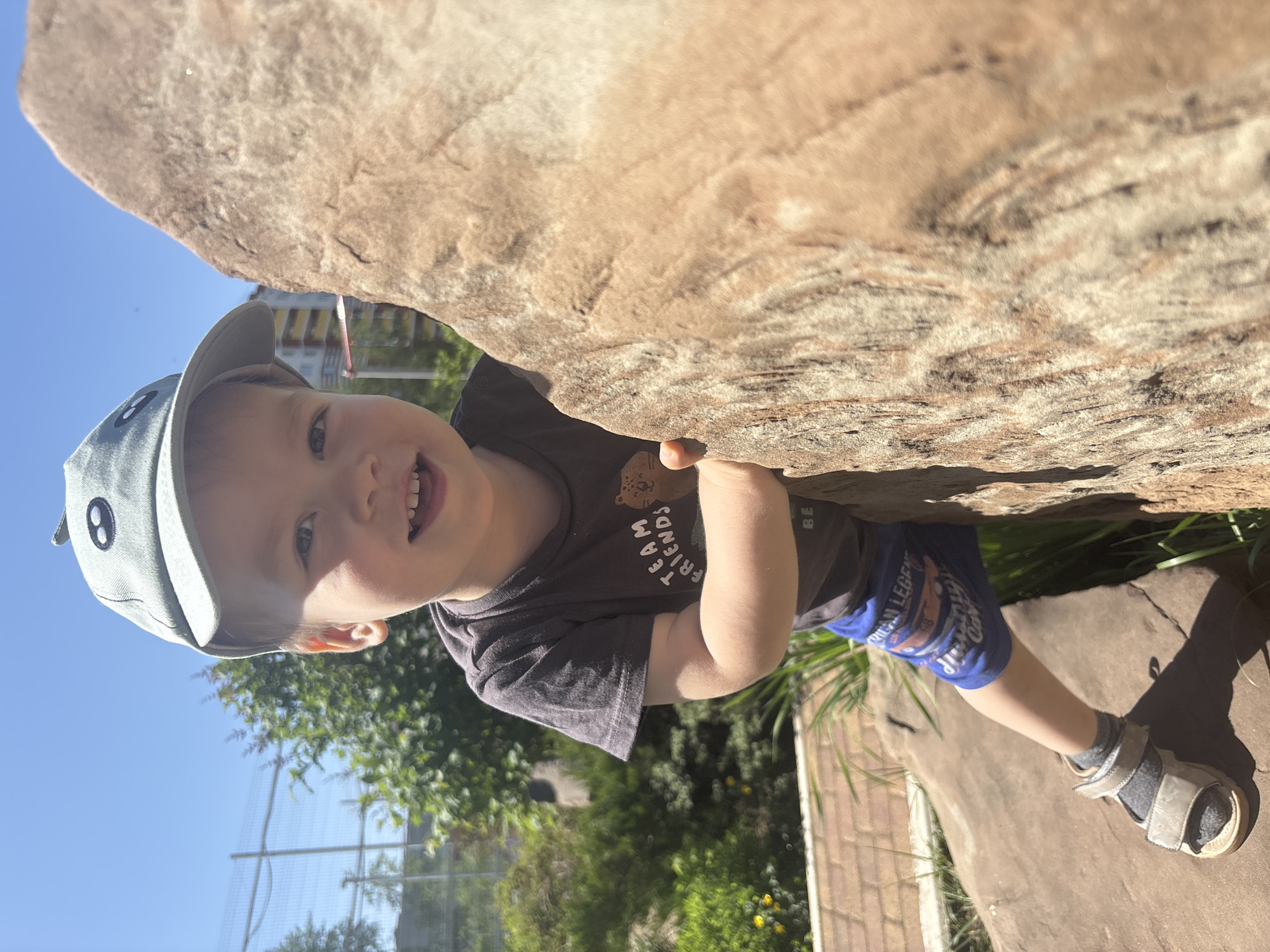 A young child wearing a blue baseball cap, a dark gray t-shirt with a bear and the words "TEAM FRIENDS" on it, blue shorts, and gray sandals stands on a reddish-brown concrete surface. He is smiling and holding onto the underside of a large, irregular, beige-colored rock. In the background, there is a green garden with a wire fence, and a clear blue sky is visible. The child is looking directly at the camera. There is no evidence to suggest this is in Izhevsk, Russia.