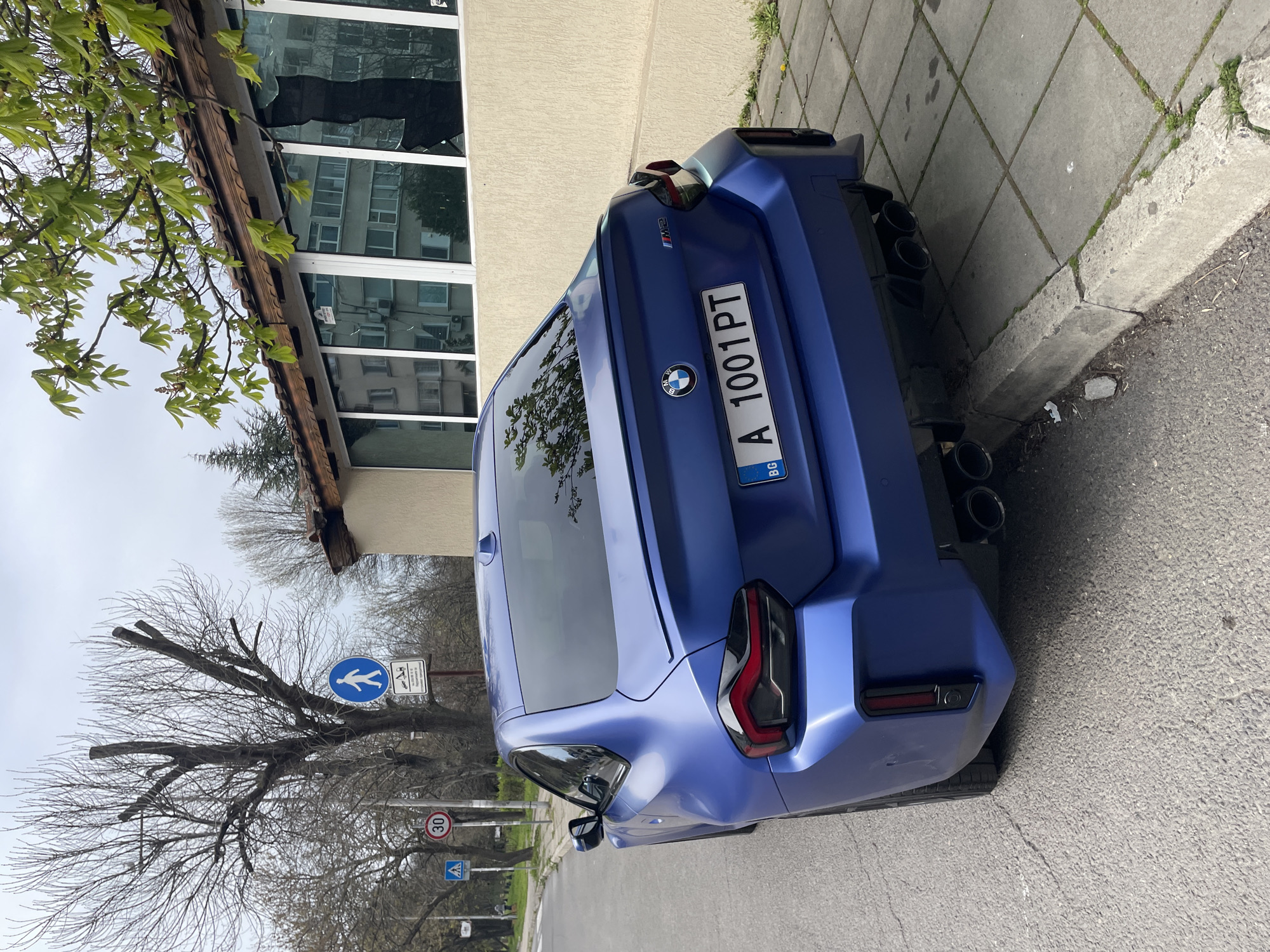 The image shows the rear of a blue BMW M2 coupe parked on a street in Burgas, Bulgaria. The car is angled towards the right, with its license plate "A 1001 PT" clearly visible. The Bulgarian flag is on the license plate. The car is parked next to a sidewalk made of grey rectangular tiles.
Behind the car is a building with large windows that reflect the surroundings. A tree with bare branches is in the background, along with street signs. One sign shows a pedestrian crossing symbol and another displays a speed limit of 30. A tree with some new green leaves visible hangs over the building.