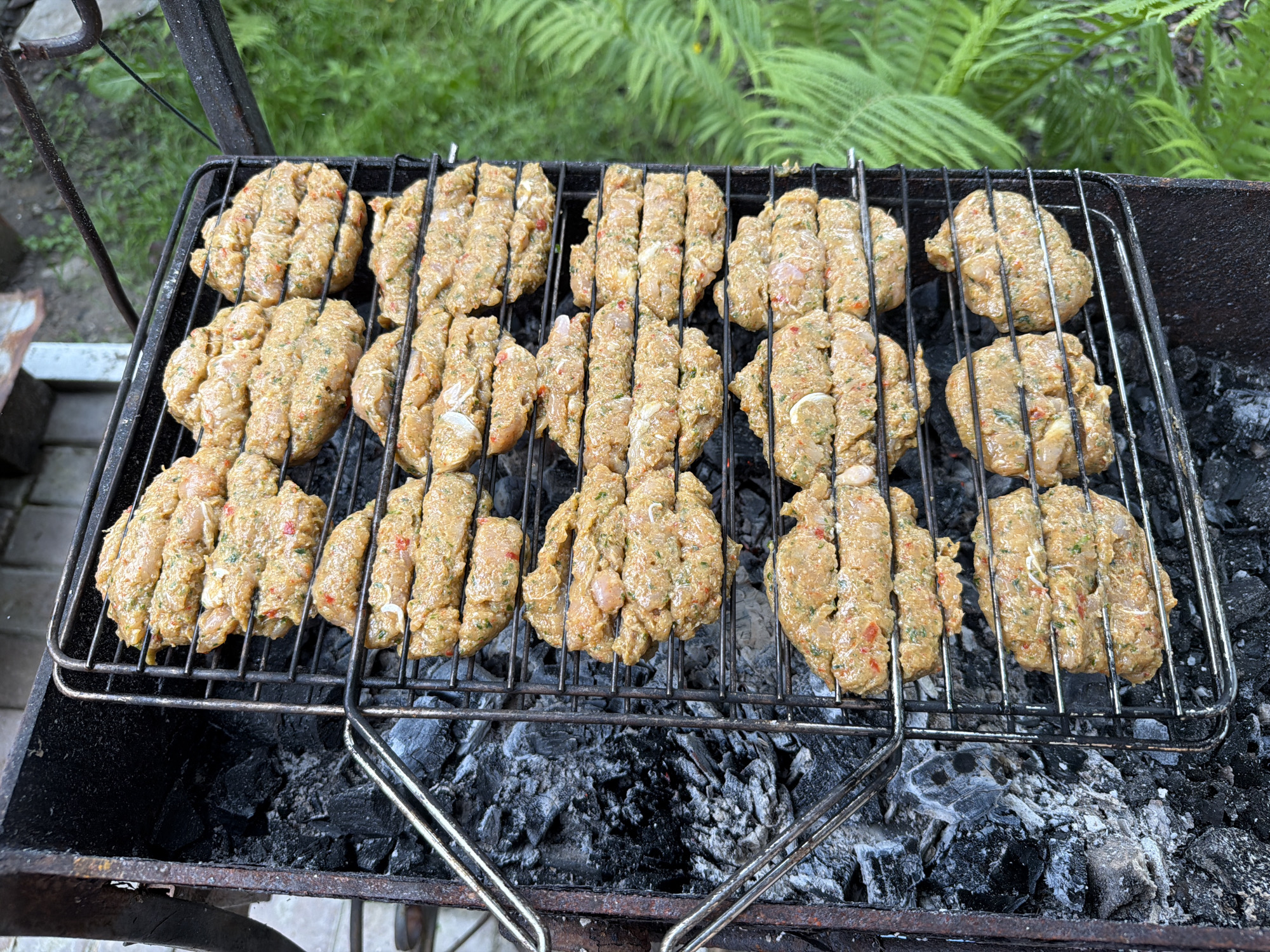 In an outdoor setting, likely a backyard in Каменка, Russia, a metal grill rack is positioned over glowing charcoal. The grill is laden with numerous raw, oval-shaped patties, appearing to be made of ground meat or a similar mixture, with visible flecks of herbs and red peppers. These patties are arranged in several rows on the wire rack. To the left of the grill, the dark, charred remains of burnt wood are visible. The background features blurred green foliage, suggesting a natural or garden environment. No people are present in the image.