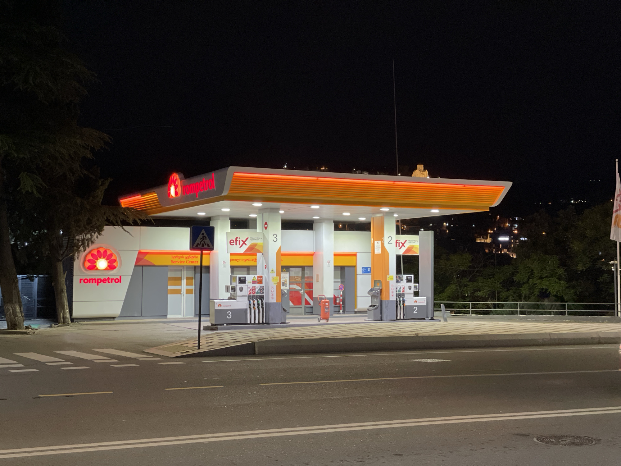 The image captures a brightly lit Rompetrol gas station at night in Tbilisi, Georgia. The scene is devoid of people and explicit activity, showing the station in a state of operational readiness.
The gas station features a modern design with a prominent orange and white color scheme. A large canopy, illuminated from below, shelters the fuel pumps. The Rompetrol logo, a stylized red floral design, is visible on the canopy's edge and on the side of the station building. Several fuel pumps are arranged beneath the canopy, each equipped with multiple hoses. The pumps are marked with numbers "3" and "2," and branding like "efix" and "fix" is also present. An orange fire extinguisher on wheels stands between two pump islands.
Behind the pumps, a service center building is visible, featuring a sign with "Service Center" in English and "სერვის ცენტრი" in Georgian. Another Georgian phrase, "ეთილი იყოს თქ," likely part of a welcome message, is also visible. The sky is dark, contrasting sharply with the station's intense artificial lighting. Distant city lights can be seen twinkling on a hillside in the background, suggesting an urban or semi-urban setting.
In the foreground, a paved road with clear white lane markings and a crosswalk is visible. Dark trees flank the left side of the gas station, with their foliage partially illuminated by the station lights. A tall, thin antenna or mast extends upwards from the gas station's right side. The overall impression is one of a functional and well-maintained establishment, standing out brightly in the nocturnal landscape.