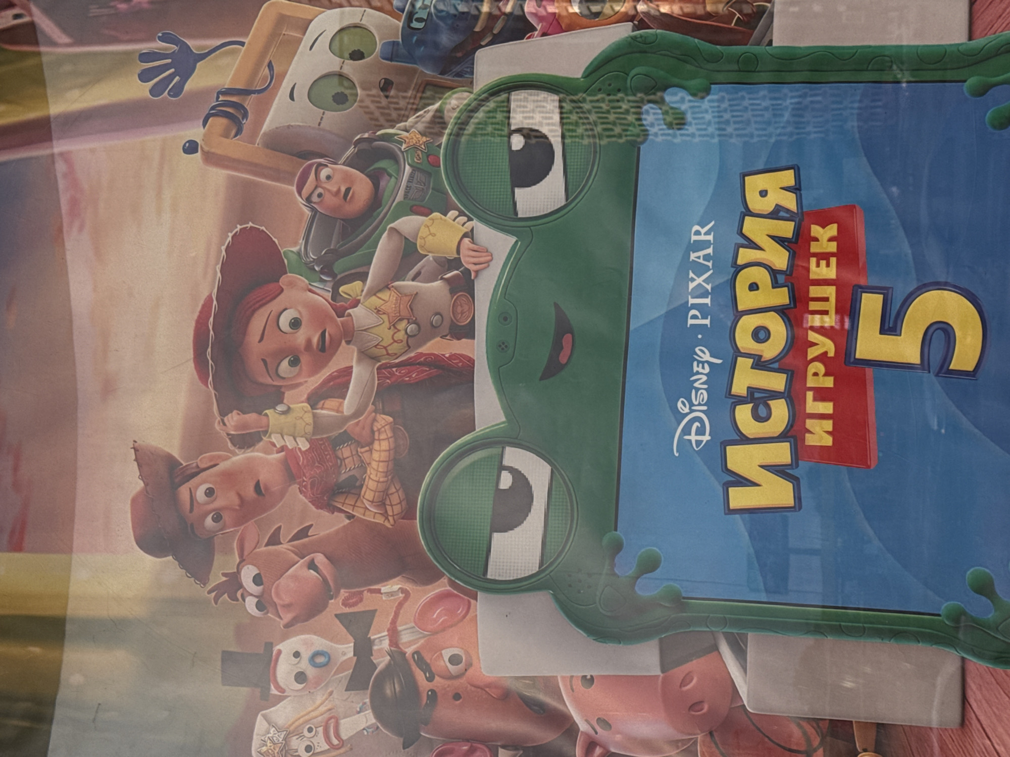 The image is a close-up, slightly angled shot of a media cover, likely a DVD or Blu-ray case, featuring characters from the Disney Pixar film "Toy Story 4". The cover is predominantly blue with a green frog-shaped border. The title, "ИСТОРИЯ ИГРУШЕК 5", is visible in large yellow letters, with "DISNEY PIXAR" also present.
The characters depicted are from the "Toy Story" franchise. In the foreground, Buzz Lightyear is visible on the left, with his characteristic space suit and determined expression. To his right is Jessie, the cowgirl doll, with her red hat and determined look. Woody, the cowboy doll, is partially visible in the lower left, recognizable by his cowboy hat and outfit. Other toy characters, including Mr. and Mrs. Potato Head, and Forky, are also present, though some are partially obscured.
The overall mood conveyed is playful and adventurous, typical of the "Toy Story" series. The image seems to be a promotional item or packaging for the film. The setting is not explicitly shown, but the cartoon nature of the characters suggests a fantasy or animated world. The lighting is bright, and there are no discernible weather cues. The location context of Krasnogorsk, Russia, implies this is a product available or marketed in that region, indicated by the Cyrillic text on the cover.