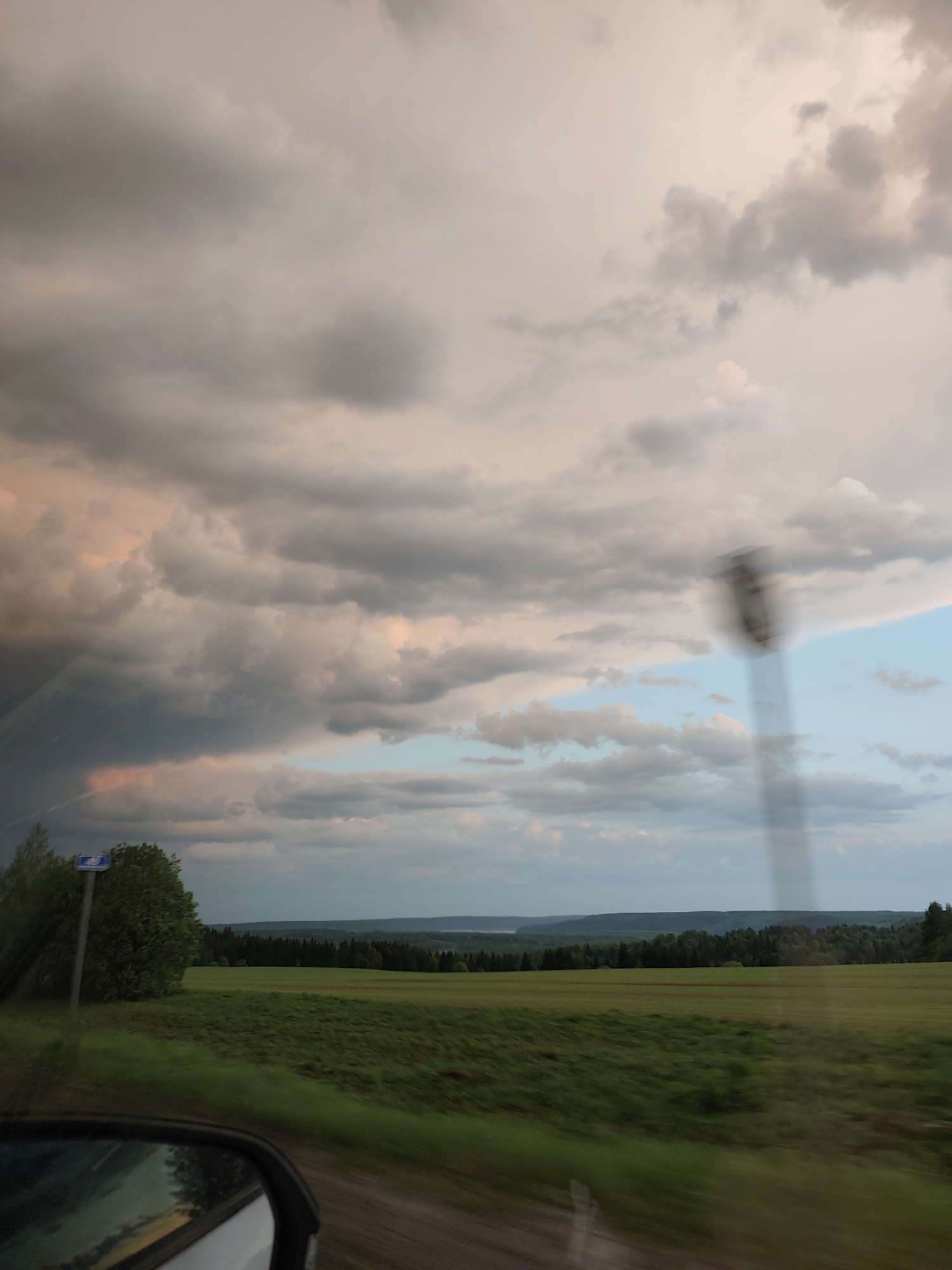 The image shows a landscape view through a vehicle window. The foreground features a green field and the side mirror of a vehicle. Further away, a dense line of trees defines the horizon. Above, the sky is filled with layered clouds, ranging from dark gray to lighter, almost pinkish hues. A blurred signpost stands along the road. Based on the location of Verkhnechusovskie Gorodki, Russia, it is most likely in a rural area. There are no people visible. The action is likely the vehicle traveling along a road.