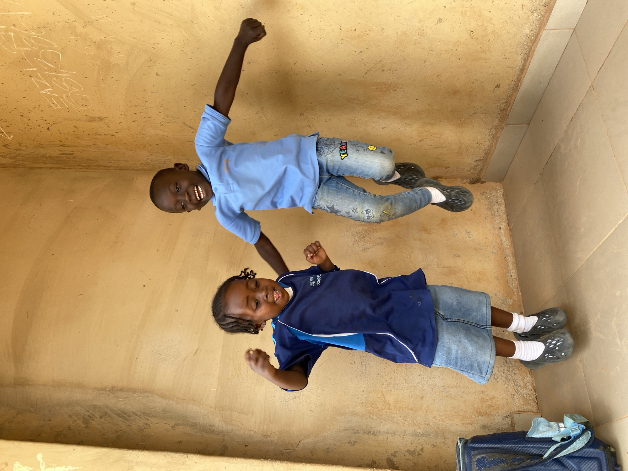 Two children are visible in an indoor location in Kuru Dungus, Nigeria. The background consists of a beige wall with some writing on it. An older child wearing a blue shirt and jeans with cartoon patches lies on their back and raises their right arm in the air with a clenched fist, smiling. A younger child wearing a navy and blue school shirt and jean skirt with white socks and black shoes lies on their back with their arms raised and clenched. A blue piece of luggage is in the bottom right corner of the image.