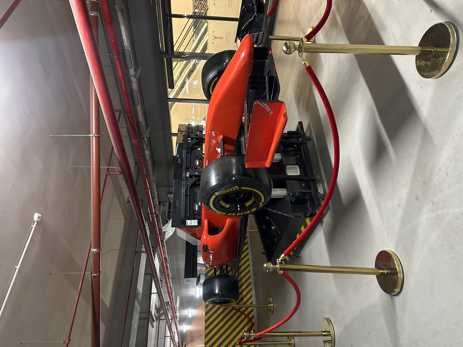 The scene depicts an unoccupied, bright red Formula 1-style racing simulator located within a commercial indoor environment, specifically identified as Tashkent City Mall in Tashkent, Uzbekistan. The simulator, featuring black tires with yellow "Pirelli" lettering and black rims, rests on an elevated black mechanical platform with visible hydraulics, suggesting motion capability. Multiple black computer monitors, branded "AOC," are mounted behind the simulator's cockpit area.
A barrier of approximately five gold-colored stanchions connected by red velvet ropes encircles the simulator on the light grey, polished concrete floor. In the background, beyond a large glass partition, multiple escalators are visible, typical of a multi-story shopping mall. A pillar through the glass clearly displays "TASHKENT CITY MALL" in white lettering. To the far left, a wall features yellow and black striped safety markings. The ceiling is an exposed industrial type, displaying numerous red and white pipes, suspended fluorescent lighting fixtures, and a white smoke detector. No individuals are present in the image, and no actions or interactions are depicted. The lighting is bright and even, casting subtle shadows from the stanchions and simulator.