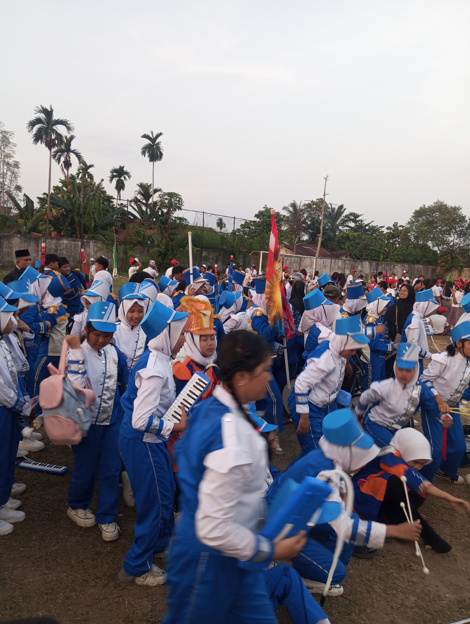 A group of young people in marching band uniforms are gathered outdoors in a grassy area. The uniforms are primarily blue and white with silver accents. Many of the individuals are wearing blue hats, some of which have a conical shape. Several people are holding musical instruments, including what appears to be a melodica and drumsticks. One individual in the foreground is carrying a light pink backpack. In the background, there is a fence, some palm trees, and buildings. The sky is overcast. The scene is consistent with a marching band practice or preparation for a performance in Candirejo, Indonesia, given the presence of palm trees and the architectural style of the background structures. The lighting suggests it is either early morning or late afternoon.