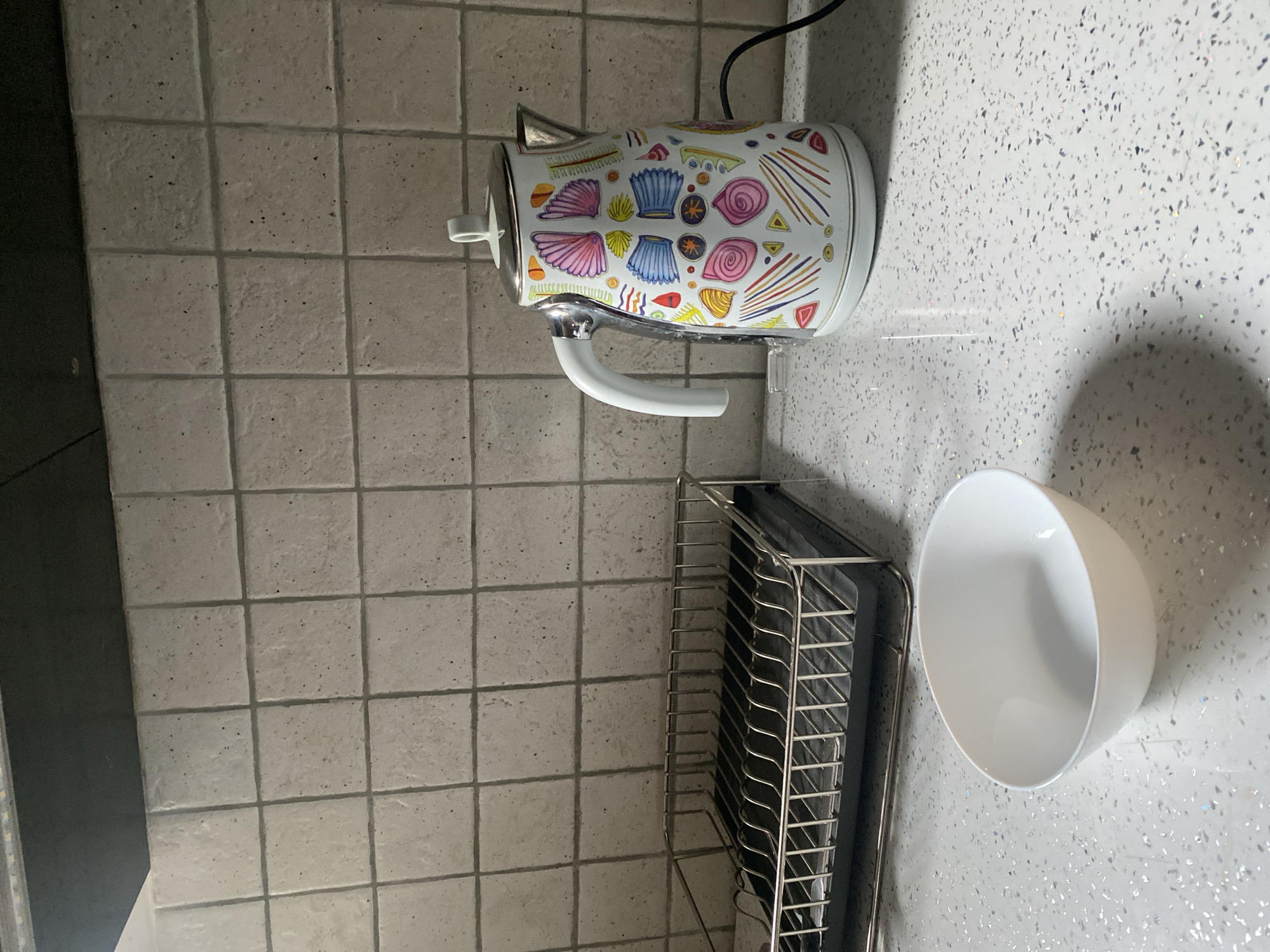A white kettle with a colorful, abstract pattern is situated on a speckled white countertop. The kettle has a silver lid and handle. To the left of the kettle is a chrome dish drying rack. In the foreground, a white bowl is placed on the countertop. The background consists of light gray square tiles. The scene appears to be a kitchen counter.