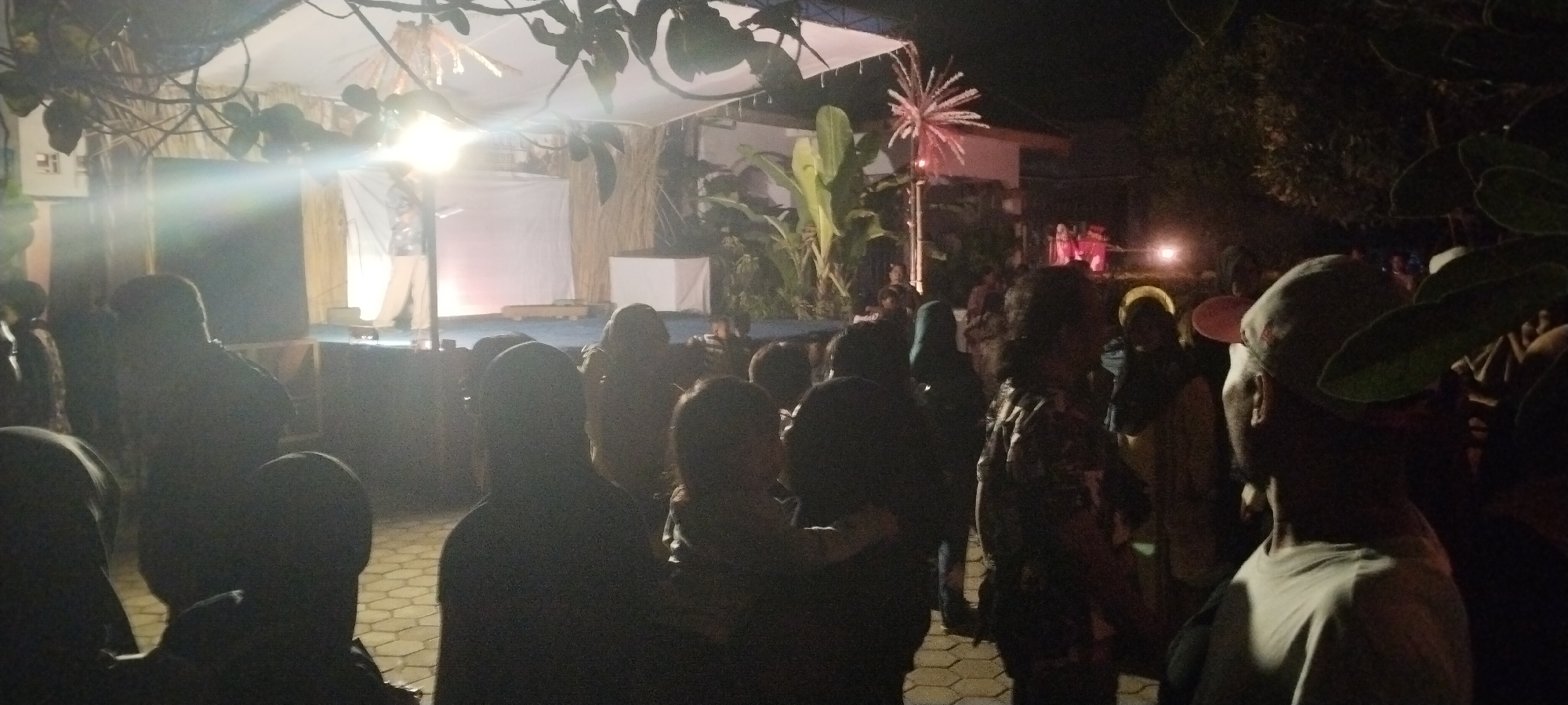 The image depicts an outdoor evening gathering, likely a performance or cultural event, in the city of Keting, Indonesia. A raised stage area is illuminated, with a person standing on it, possibly an entertainer or speaker. Behind the stage, there are large potted plants and some decorative elements, including a spiky red light fixture. The audience is gathered in front of the stage, with many individuals silhouetted against the light. The scene suggests a community event, possibly a festival or celebration. The ground appears to be paved with hexagonal tiles.