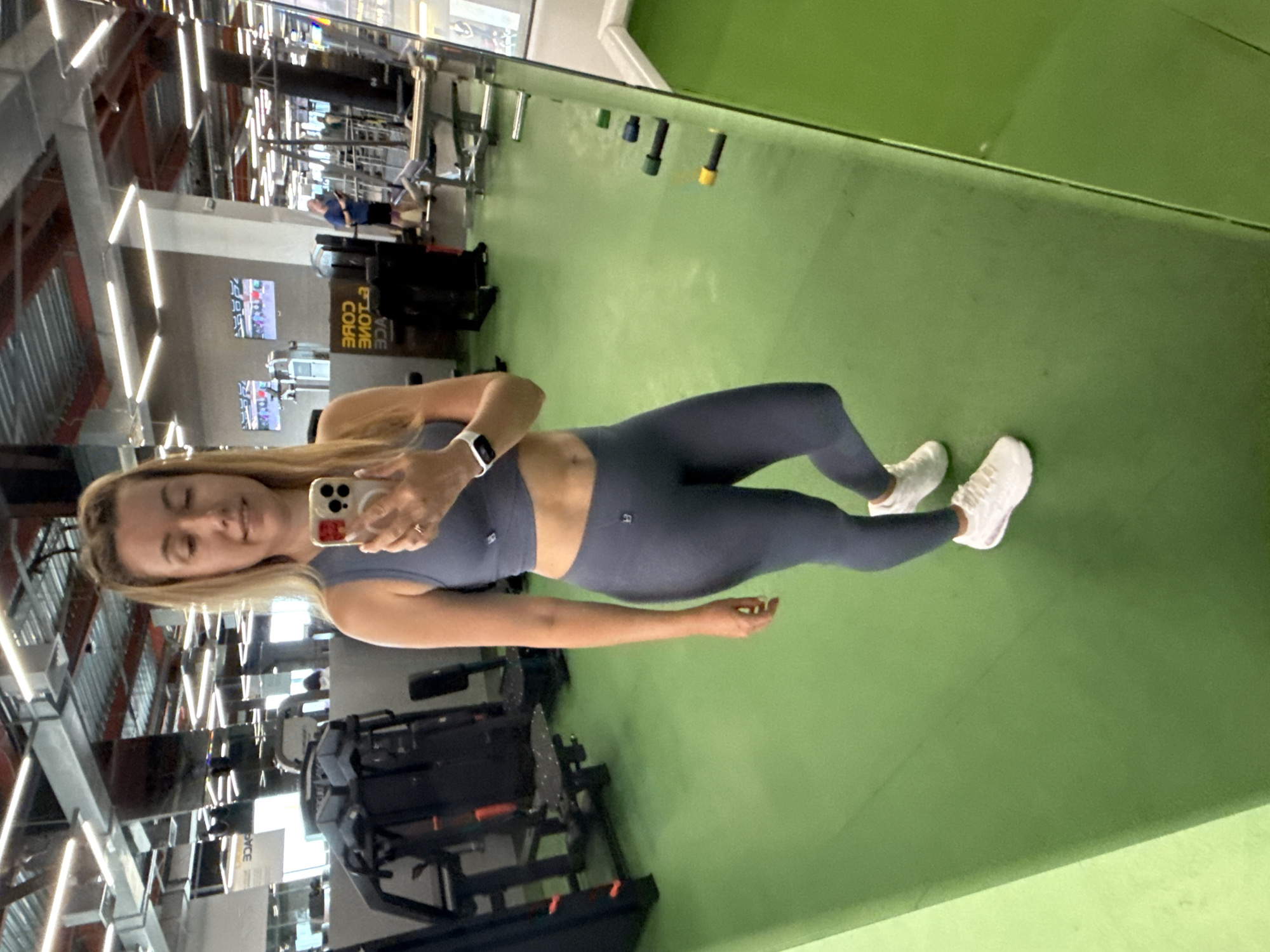 A woman stands in a gym, taking a mirror selfie. She has blonde hair pulled up in a ponytail and is wearing a gray sports bra and matching leggings, along with white sneakers. Her smartwatch is visible on her left wrist. The gym has green flooring and various exercise machines and equipment in the background. The overall lighting suggests an indoor environment.