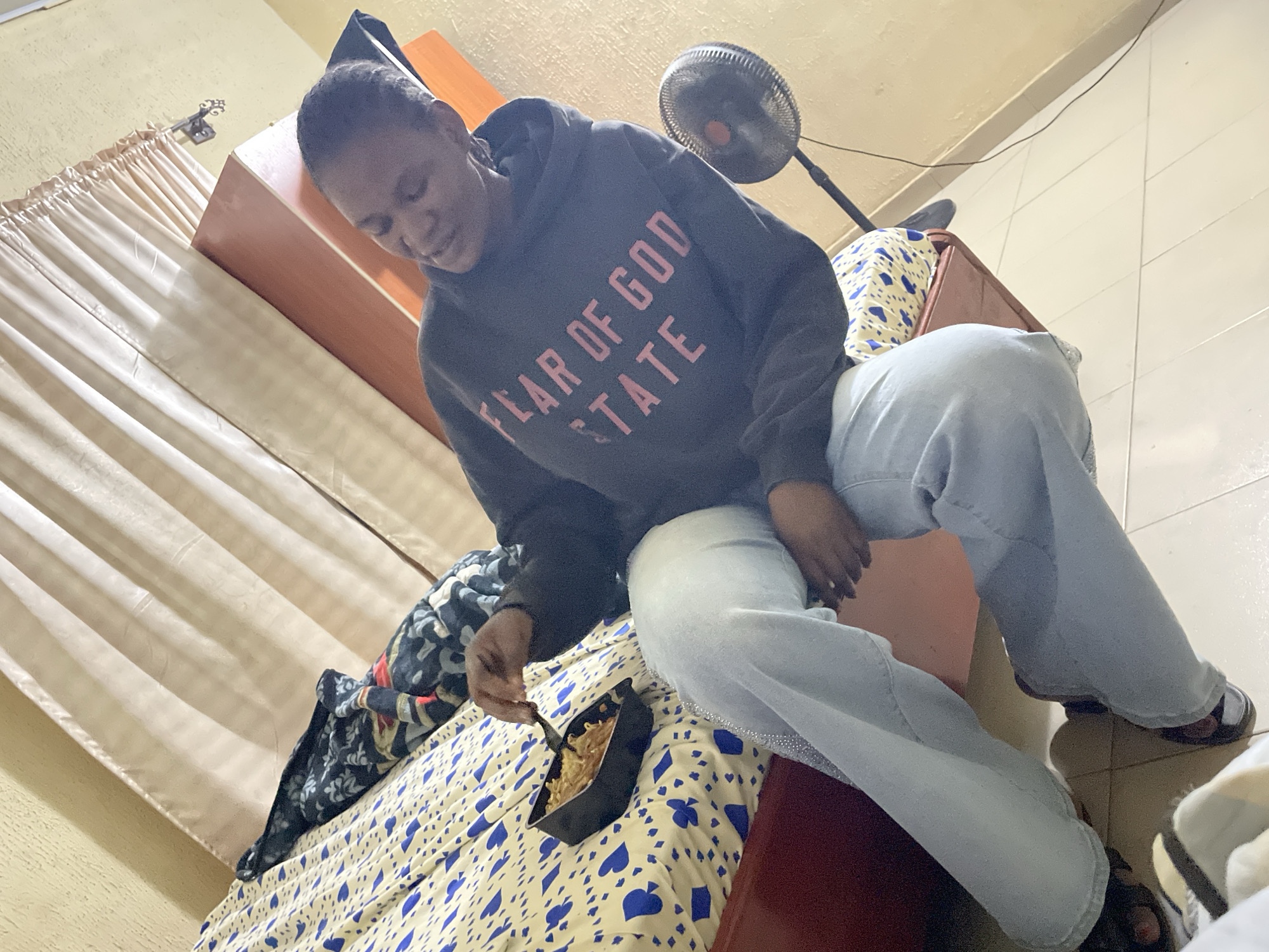 A person is sitting on a bed in a room. The person is wearing a gray hoodie with the words "FEAR OF GOD STATE" in pink lettering and light blue jeans. They are eating from a black container with a fork. The bed has a patterned blue and white bedspread. The room contains a wardrobe or dresser, a curtain, and a fan. The specific location within the city of Jos, Nigeria cannot be determined from the visual information provided.
