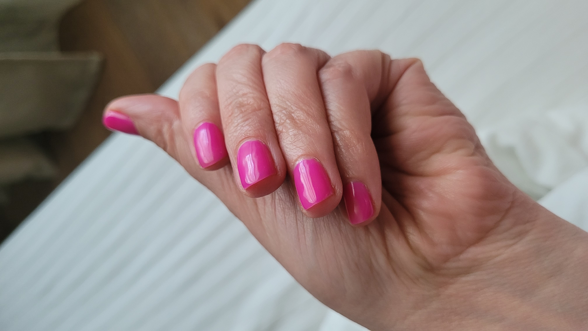 The image shows a close-up of a person's left hand with bright pink nail polish on all five fingernails. The hand is positioned with the palm facing upwards and the fingers slightly curled inwards. The skin on the hand appears fair and shows some natural creases. The nails are short and have a glossy finish. The background is out of focus but appears to be a white, textured surface, possibly bedding or a towel, with faint diagonal lines. To the left of the hand, there is a blurred area that seems to be furniture or a wall with a warmer, neutral tone. The lighting suggests an indoor setting, possibly during the day, casting soft shadows. There is no visible text in the image. The overall impression is one of personal grooming or a moment of relaxation.