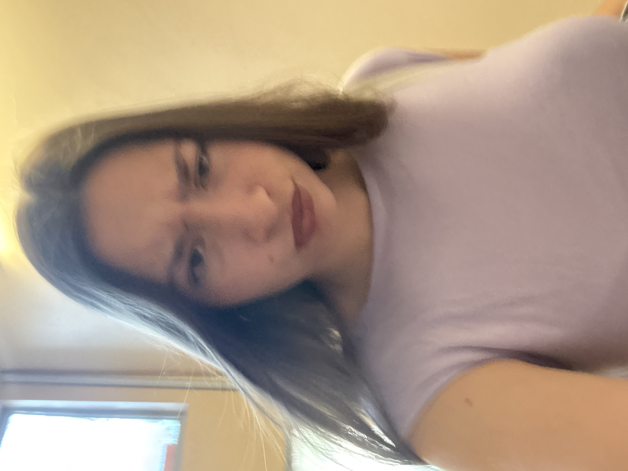 A medium shot captures a young woman from the chest up, facing the camera with a displeased expression. Her eyebrows are furrowed, and her lips are pursed. She has long, dark hair that frames her face, with some strands falling forward. She is wearing a light purple, short-sleeved shirt. The background is a blurred warm yellow wall with a bright, indistinct window on the left side, suggesting an indoor setting, possibly an apartment or office in Sofia, Bulgaria. The lighting is warm and diffused.