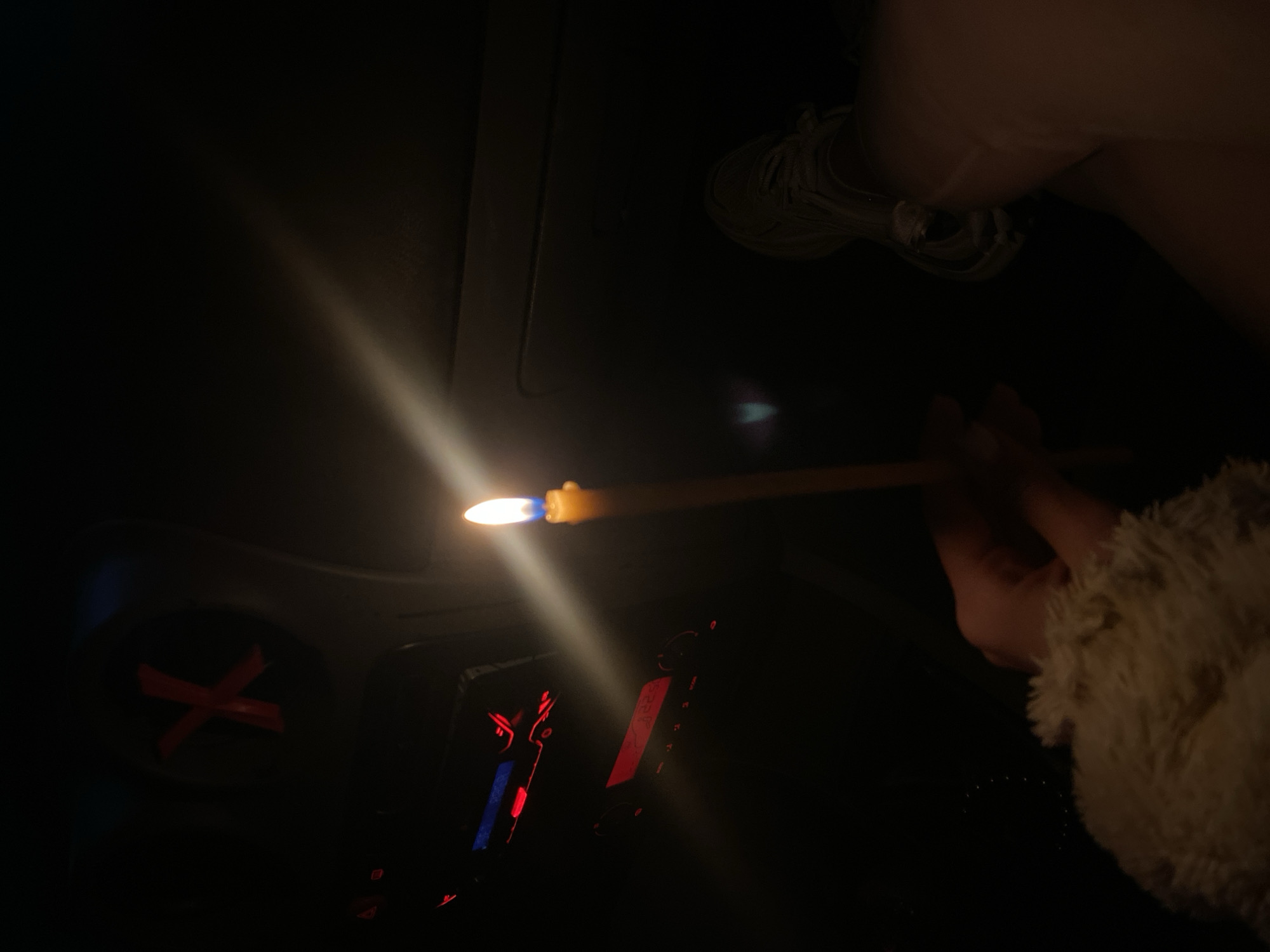 A person is holding a lit candle in a dark interior, possibly a car. A dashboard with red lights and a red "X" taped to an area above the dashboard are visible. The location is unknown, and there is no way to determine if it is in Blagoevgrad, Bulgaria.