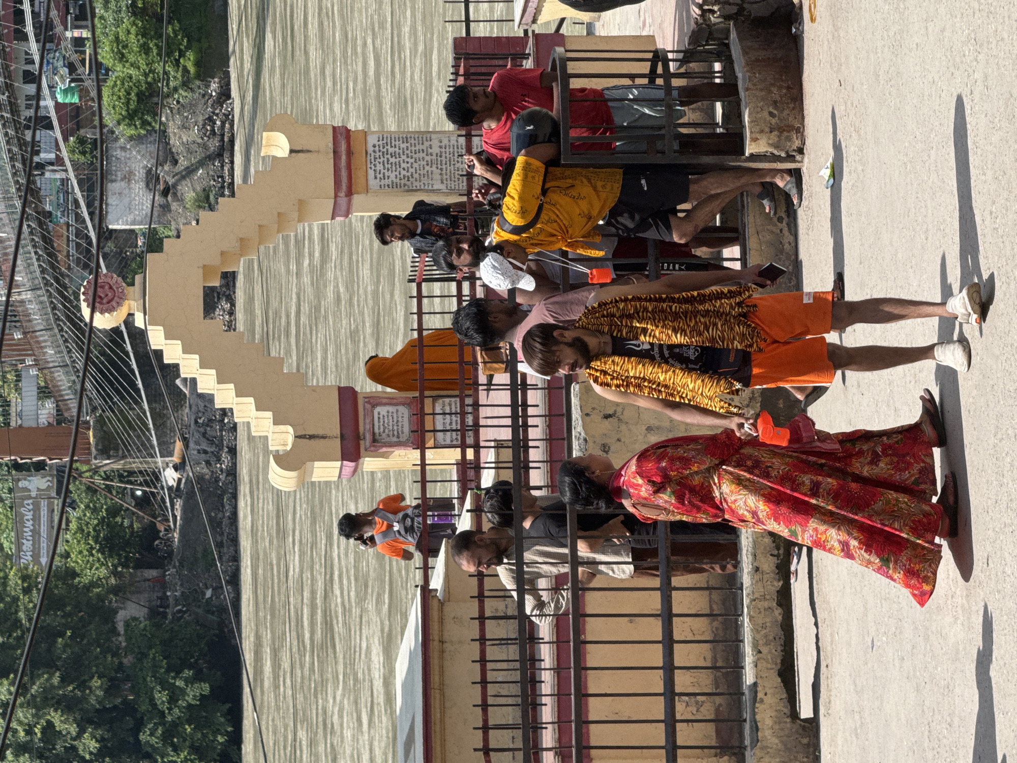 In Rishikesh, India, a group of people are gathered near the Ganges River. In the foreground, a man in orange shorts and a tiger-print garment stands, holding a phone. To his left, a woman in a red floral dress walks away, holding an orange object and a pink cloth. Behind them, a black metal fence separates the onlookers from a stone wall and a walkway. Further back, several men stand behind the fence, some talking and one holding a black helmet. The backdrop features a wide, muddy river, a large arched structure with a decorative element at its peak, and lush green trees on the far bank. A sign with Hindi text is visible on a pillar near the arched structure. The scene is brightly lit by sunlight, casting distinct shadows on the ground.