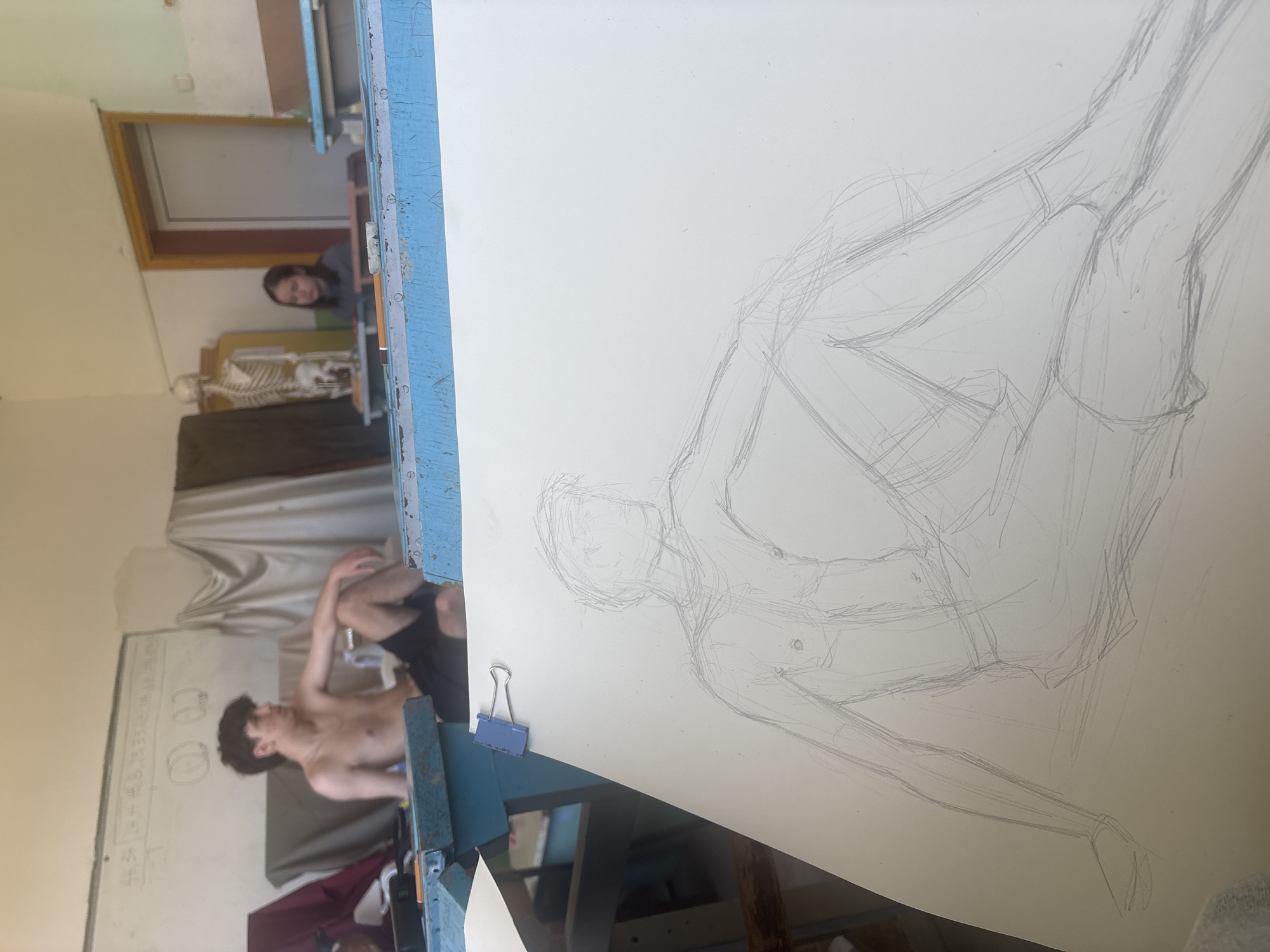 The image shows a figure drawing class in a room in Troyan, Bulgaria. In the background to the left, a partially nude male model with short dark hair sits with his right leg bent and foot resting on his left knee, his right arm resting on his knee and his left arm slightly raised. He wears dark shorts. Behind him is a whiteboard covered in writing, and a gray curtain or backdrop. In the background towards the center, a skeleton model is visible on a table or shelf. A female observer with dark hair looks out from a doorway behind the skeleton. In the foreground, a drawing of the model on a large sheet of paper is clipped to a blue easel. The drawing is a sketch, showing the model's pose. The room has light walls and appears to be a studio or classroom.