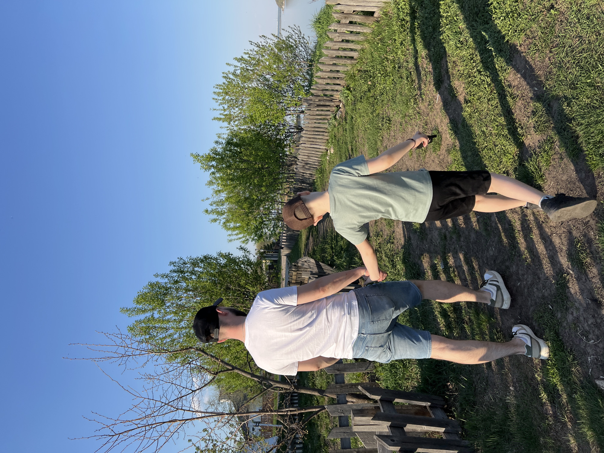 Two people, an adult and a child, stand on a grassy path next to a wooden fence under a clear blue sky. The adult, wearing a white t-shirt, denim shorts, and a black baseball cap, appears to be holding hands with the child. The child wears a light green t-shirt, black shorts, a brown hat, and points an object in their right hand away from themselves, out of the scene. The location is outdoors, possibly near a park or countryside area, with trees and vegetation visible in the background. The wooden fence runs along the edge of the path. The city is Sviyazhsk, Russia.