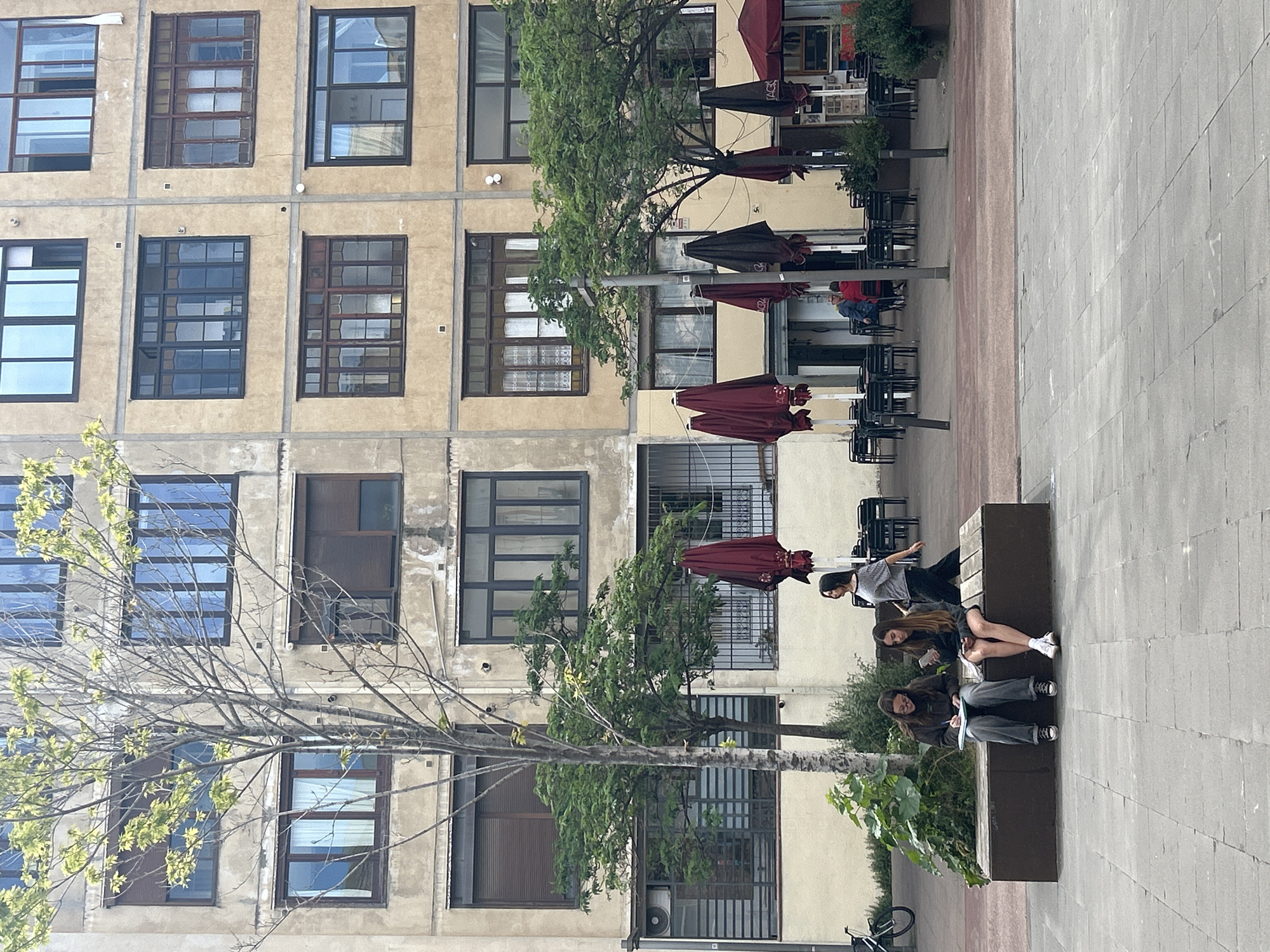 The image depicts a street scene in Barcelona, Spain. A multi-story building with numerous rectangular windows is in the background. A row of trees lines the sidewalk, partially obscuring the building. Several outdoor seating areas with tables, chairs, and maroon umbrellas are visible along the building's facade. In the foreground, three individuals are seated on a park bench. One appears to be writing or drawing in a notebook. The ground is paved with rectangular stone blocks. A bicycle is parked near the bench.
