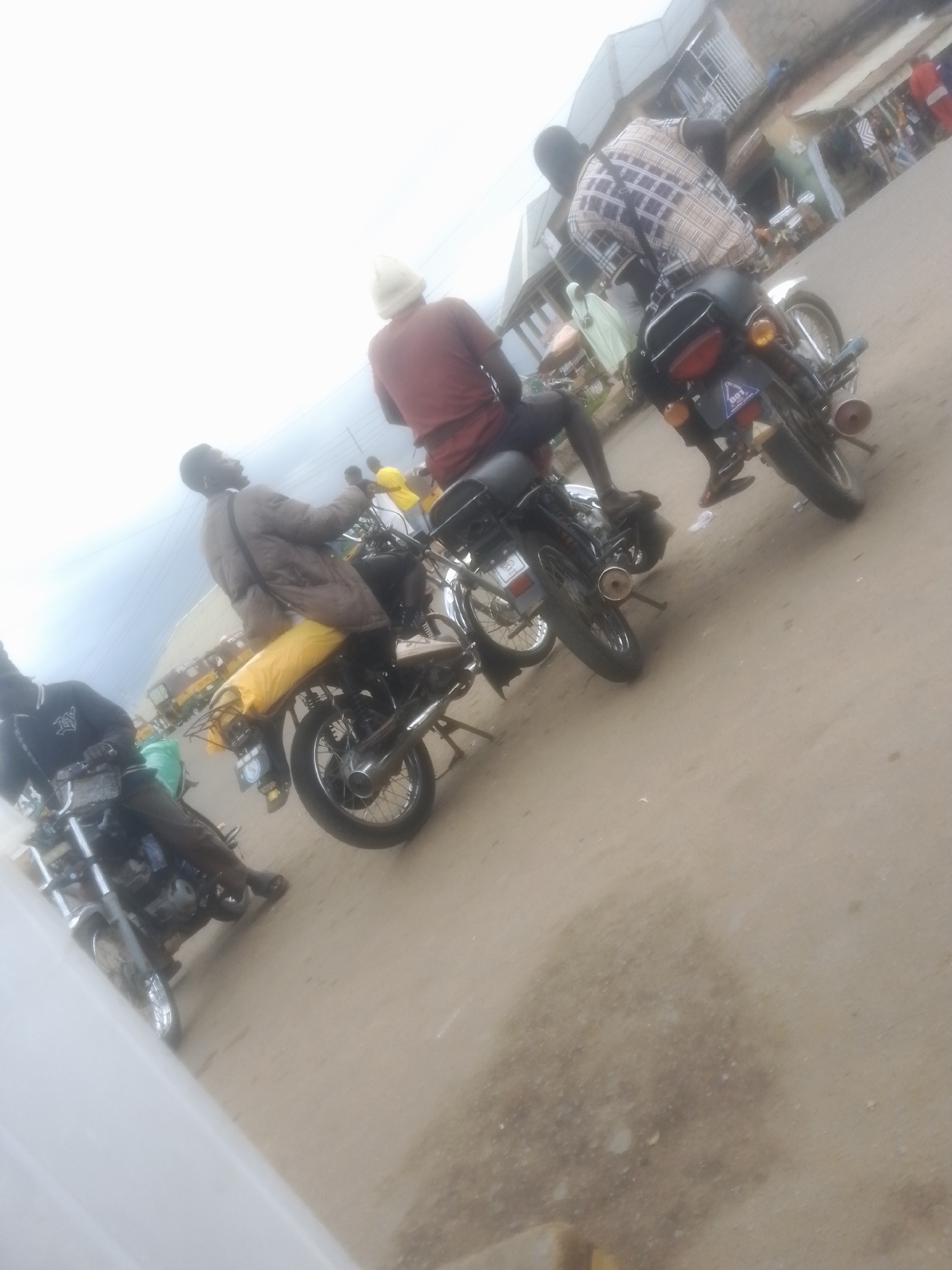 The image captures a street scene with several motorcycles and individuals. In the foreground, a person is seated on a motorcycle, wearing a dark blue shirt and dark pants. Behind them, another motorcycle is visible with a rider wearing a light brown jacket and yellow trousers. Further in the background, two more motorcycles are present. One has a rider in a plaid shirt and another individual in a red shirt and white head covering is seated behind them. The environment appears to be an outdoor urban setting with buildings and power lines visible. The ground is paved. Based on visual cues like architecture and the presence of people, this scene is likely located in Gwong, Nigeria.