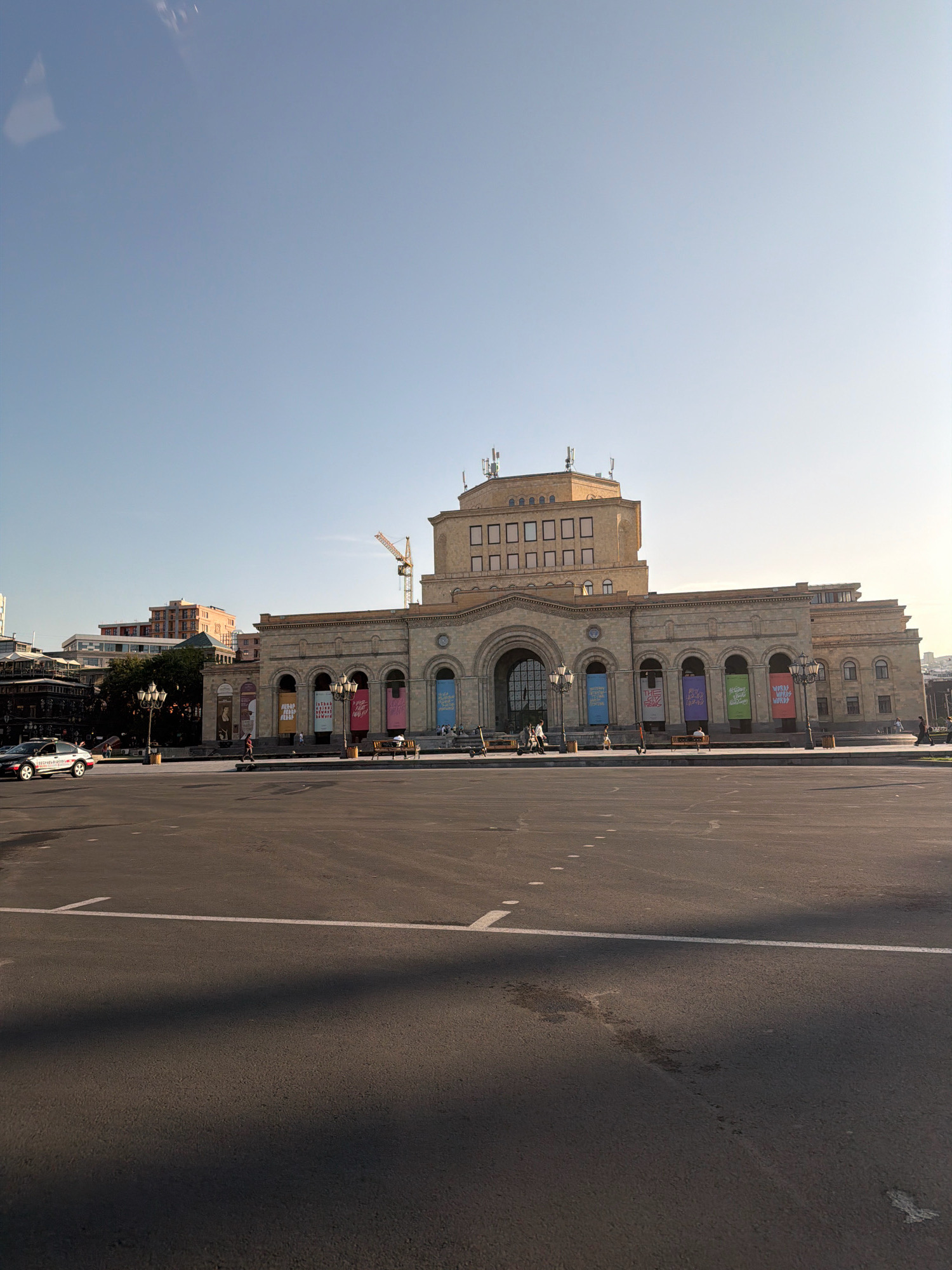 The image depicts a wide, paved plaza in front of a large, classical-style building in Yerevan, Armenia. The building, likely a museum or cultural center, is made of light-colored stone and features a prominent central entrance with a large arched doorway. Above the entrance, the building has a stacked, tiered design with rows of windows. Several colorful banners are hung along the facade of the building, some displaying text.
In the foreground, the plaza is mostly empty, with a few white lines marking parking or pedestrian areas. The asphalt is somewhat worn, with some darker patches and scuff marks. A shadow from an unseen object stretches across the foreground.
On the left side of the plaza, a white taxi is parked near the curb. Several people are visible walking on the plaza and near the building. Some electric scooters are also present. In the background, there are other buildings, including a taller modern structure with a light orange facade, and a construction crane is visible behind the main building. The sky is a clear, pale blue with a hint of sunlight, suggesting it is daytime. The overall scene is one of a public space in an urban environment, with a focus on the grand architecture of the central building.