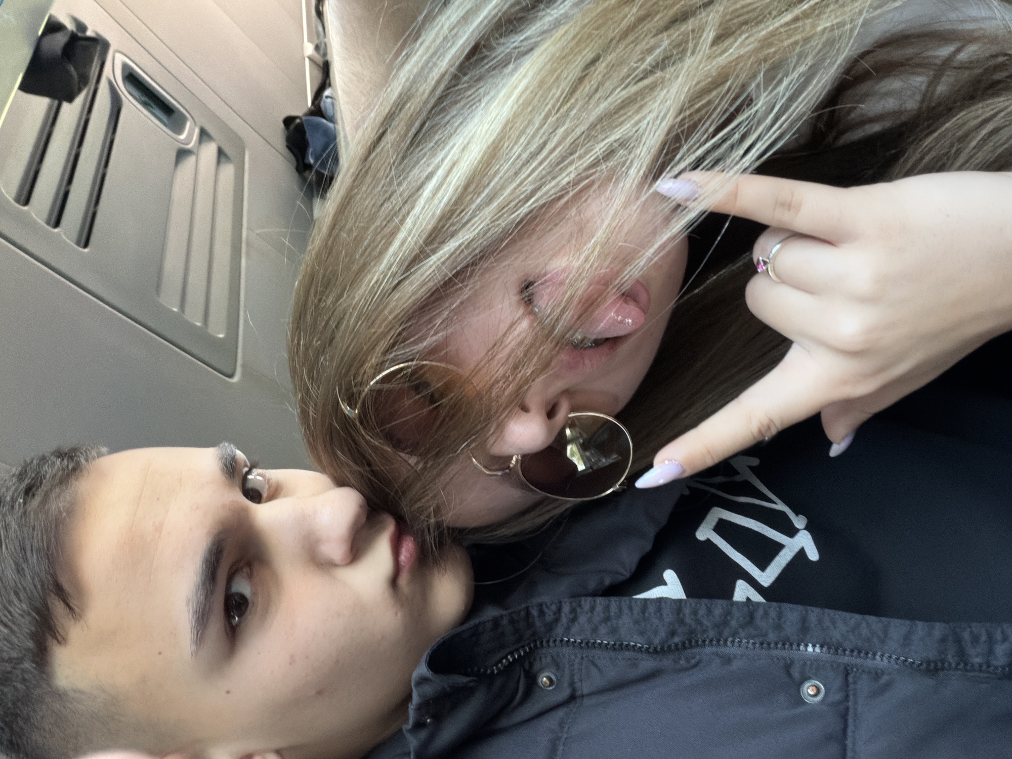 The image depicts two individuals, likely a male and female, in a close, possibly intimate setting. The female, with blonde hair and sunglasses, is making a gesture with her hand and sticking out her tongue. She is wearing a ring on her finger and has elongated manicured nails. The male, with dark hair, is looking towards the female. They are both wearing dark clothing, with the female's shirt displaying a white graphic. The background suggests the interior of a vehicle, possibly a bus or a train, based on the presence of a storage compartment. The location is not evident from the image itself. There is no information present in the picture to indicate they are in Kapitan Andreevo, Bulgaria.