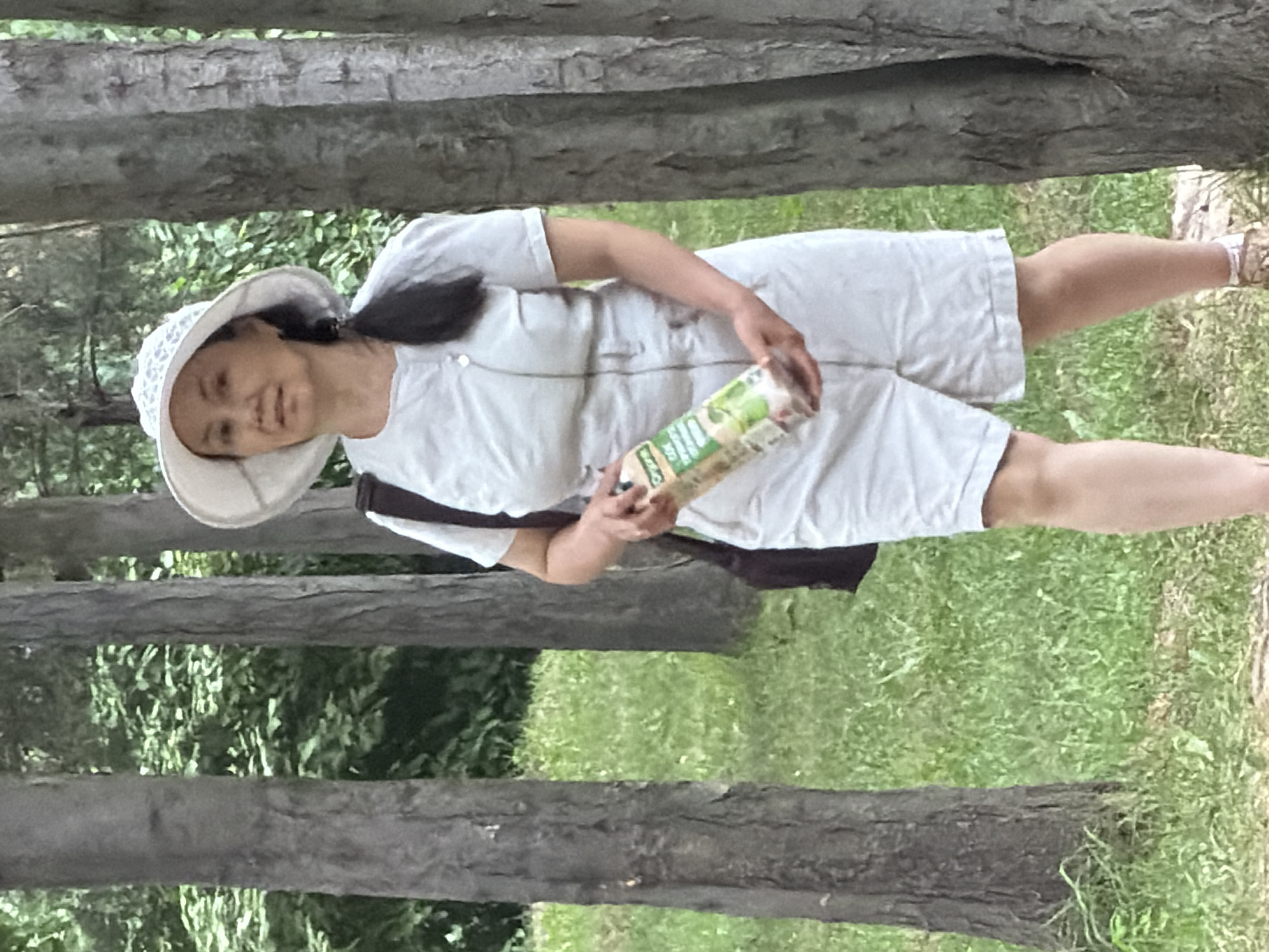 A medium shot captures a woman in a light-colored outfit and a wide-brimmed hat standing outdoors among trees. She is holding a juice carton in her right hand. The woman appears to be middle-aged, with dark hair pulled back into a ponytail. Her outfit consists of a short-sleeved top and shorts. The background is a blur of green foliage and tree trunks. The lighting suggests it is daytime, likely with some overcast. The scene is peaceful and natural, hinting at a casual outing or walk in a park.