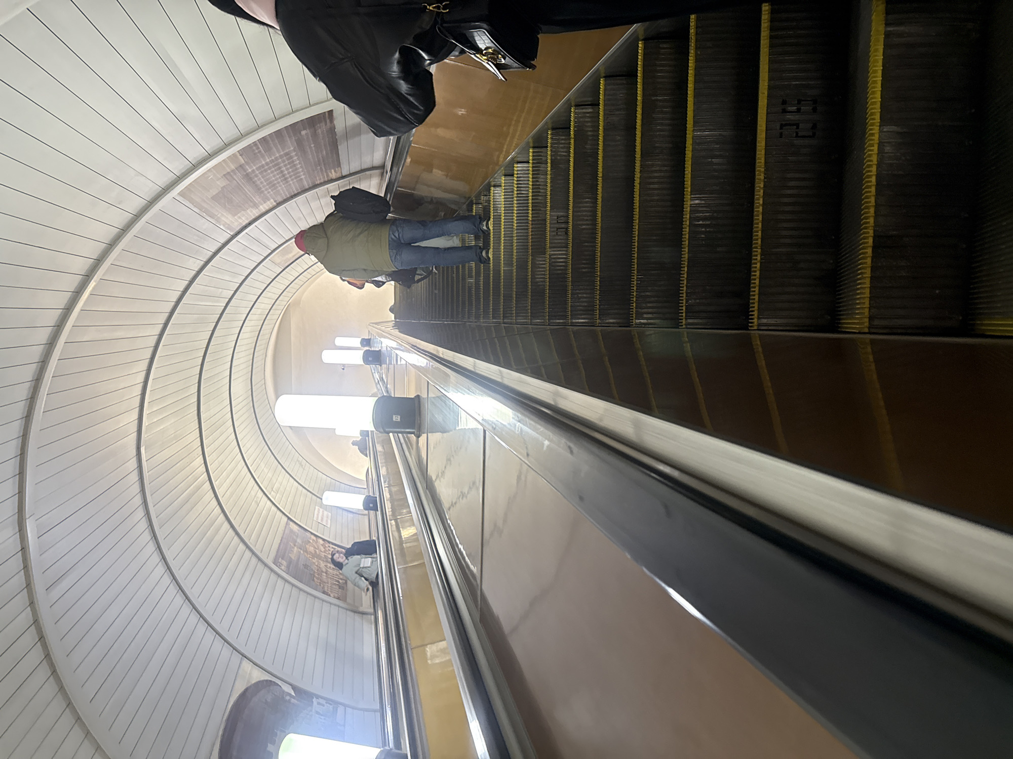 The image captures a view from the top of a long escalator in a circular tunnel, most likely in the Yekaterinburg Metro system, Russia.
Several people are visible riding the escalator. The person closest to the top is wearing a dark-colored coat and holding a purse. Further down, there is a person wearing a green jacket, blue jeans, and a red hat, also riding the escalator. Near the bottom, some more people are visible.
The environment consists of a white, curved tunnel ceiling with linear patterns. The walls of the escalator shaft are finished with light brown panels. The escalator itself has black steps with yellow markings and a silver handrail. Overhead fluorescent lights illuminate the tunnel.