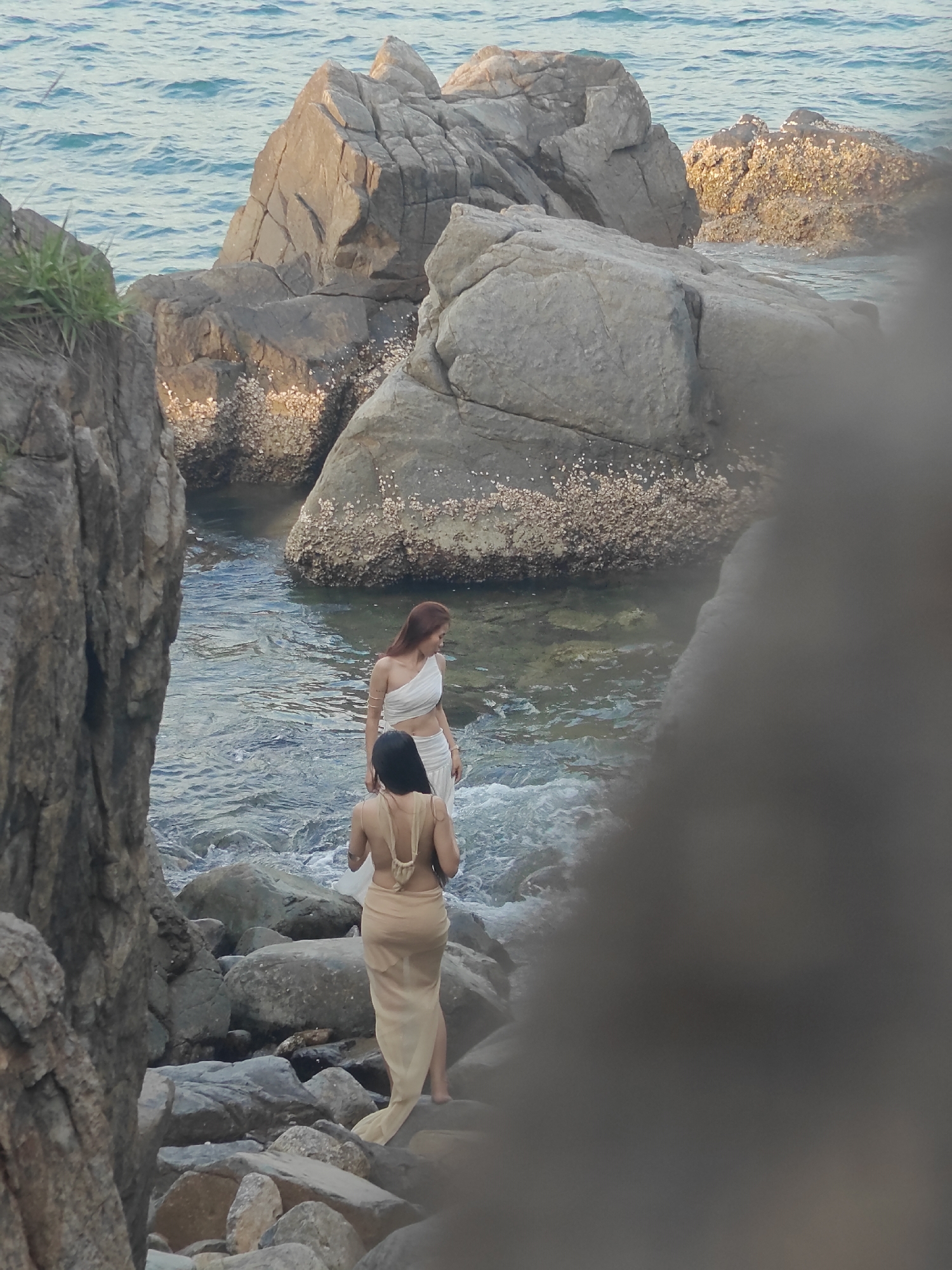 The image shows two women standing on rocks by the sea. The woman in the foreground is facing away from the camera, wearing a sheer beige sarong that drapes over her body. She has dark hair. The woman in the background is facing to her left, wearing a white one-shoulder top and a matching white skirt. She has auburn hair. The environment is rocky with large boulders and the ocean waves are visible. The lighting suggests it might be dusk or dawn. The location is identified as Vĩnh Lương, Vietnam.