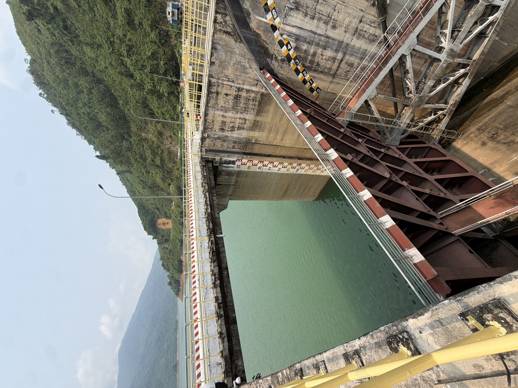 The image depicts a section of the Hồ Hòa Trung dam, located in Đà Nẵng, Vietnam. The view is from a high vantage point, likely from the dam's structure itself, looking down at the reservoir. The water in the reservoir is a greenish hue. The dam's concrete structure is visible, with various levels and support beams. A metal safety railing with yellow and white stripes runs along the edge of the dam. Markings on the concrete indicate water levels. Lush green vegetation covers the hillsides in the background. A light-blue sky is present in the upper left corner of the frame.