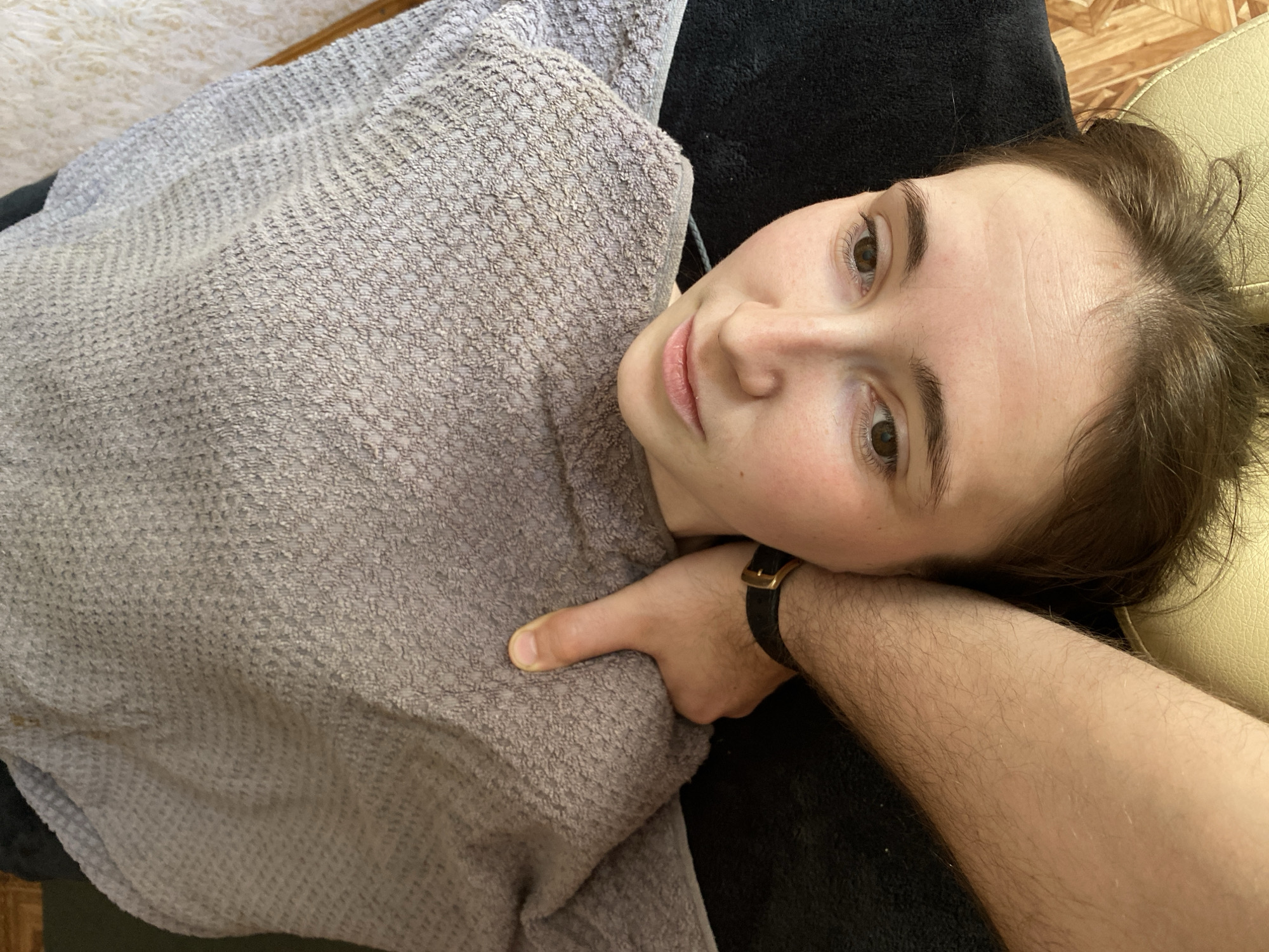In a close-up, overhead shot, a person's face, with brown eyes and light pink lips, is tilted slightly upwards, looking towards the camera. A grey textured towel is draped over their head and shoulders, obscuring part of their face and body. A hand, with a black watch on the wrist, is positioned under the chin, gently holding the head. The background consists of dark fabric and a light-colored textured surface, suggesting an indoor setting. The overall lighting is soft, creating subtle shadows on the face.