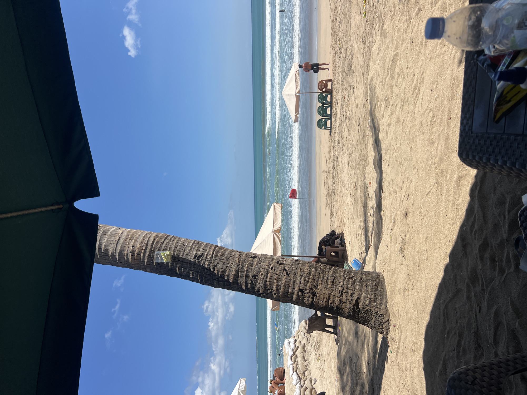 A sunny day at Kuta Beach, Indonesia, is depicted in this image. In the foreground, a large palm tree trunk dominates the left side of the frame, with a clear plastic cup containing what appears to be green fruit slices tied to it. The beach is sandy, with shadows cast by an overhead umbrella suggesting a seating area in the lower foreground.
Further back, beach chairs and umbrellas are set up on the sand. A beige umbrella is prominent, with three green plastic chairs and one orange plastic chair arranged in front of it. A man is standing near these chairs, facing the ocean. To the left, a sandbag barrier is visible, with two people partially obscured behind it, sitting on beanbag chairs.
The ocean stretches out to the horizon, with gentle waves rolling onto the shore. The water is a vibrant turquoise, contrasting with the blue sky dotted with scattered white clouds. A red flag is planted in the sand near the umbrella and chairs, likely indicating a swimming safety warning. Several people can be seen in the water, some surfing on small waves. The overall atmosphere is one of a typical beach day in a popular tourist destination.