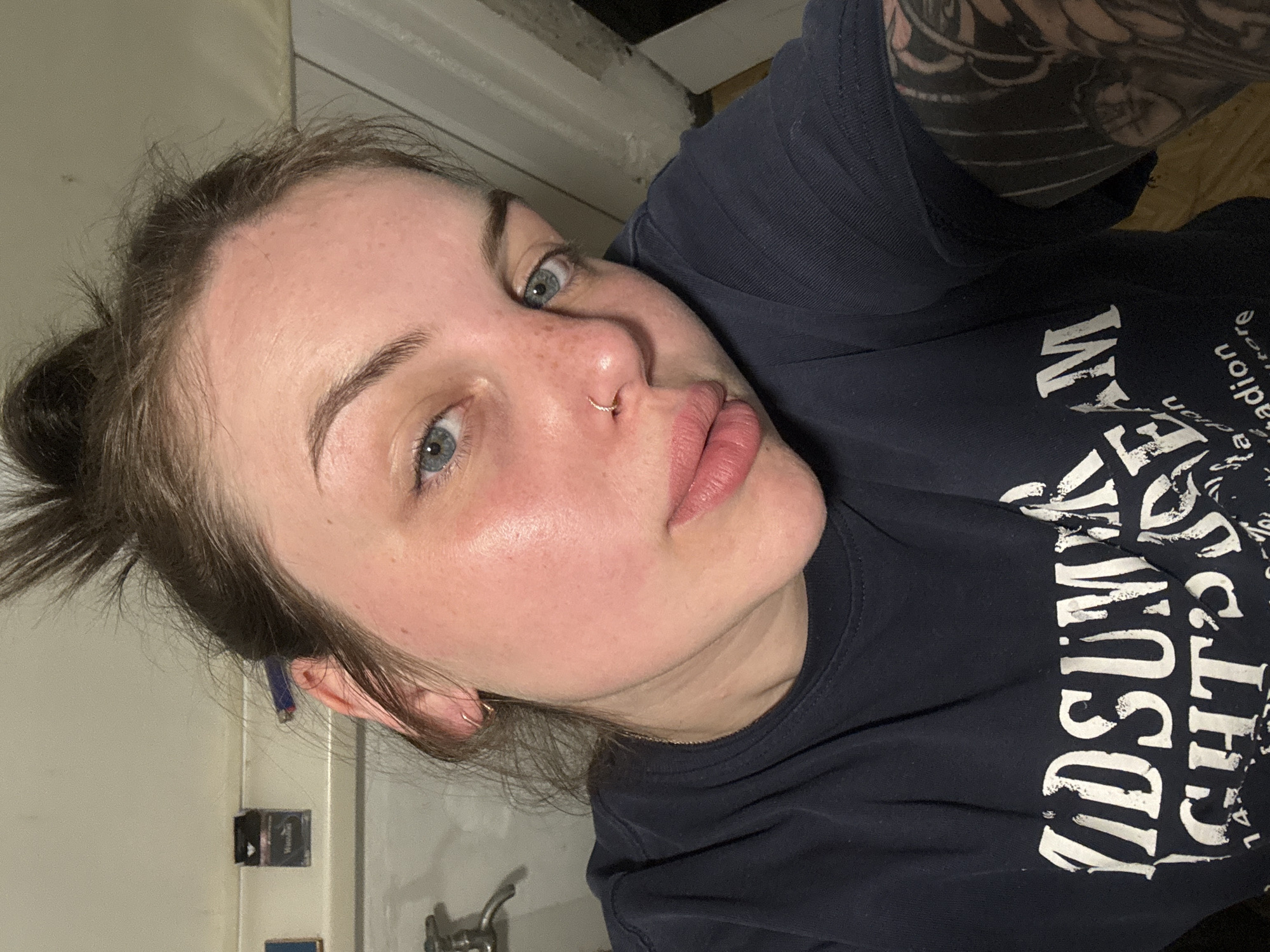 A woman with fair skin, light blue eyes, and brown hair pulled back in a bun is shown in a close-up, selfie-style photograph. She has a nose ring in her left nostril and is wearing a dark navy-blue T-shirt with white lettering. A portion of a tattoo is visible on her right arm. The background appears to be a tiled bathroom, with a sink and wall accessories visible. The location in the city of Chornomorskoe, Ukraine cannot be independently confirmed from the provided image.