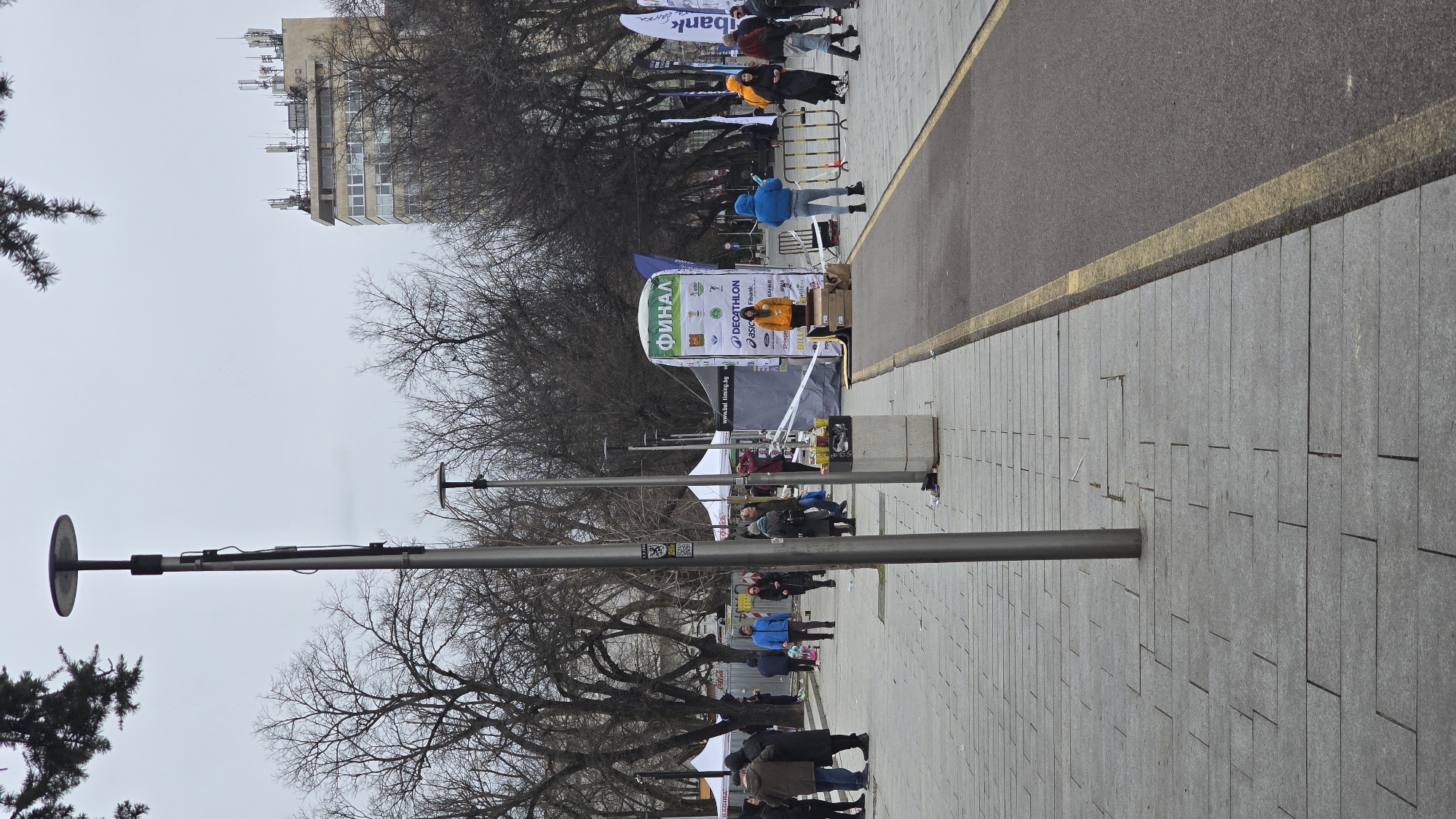 The image depicts a scene on a pedestrian walkway in Stara Zagora, Bulgaria. The ground is paved with large rectangular grey stones. A narrow asphalt path runs parallel to the paved area, separated by a yellow line. Along the walkway, there are tall metal lampposts with round, flat lamps. Bare trees line the walkway. Several people are walking or standing along the walkway. There is a banner with a white background and several logos, including "ФИНАЛ", "DECATHLON", "ASICS", and "Fibank". A woman in an orange jacket is standing behind a table near the banner. A building is visible in the background, with a facade consisting of repeating rectangular windows. The sky is overcast.