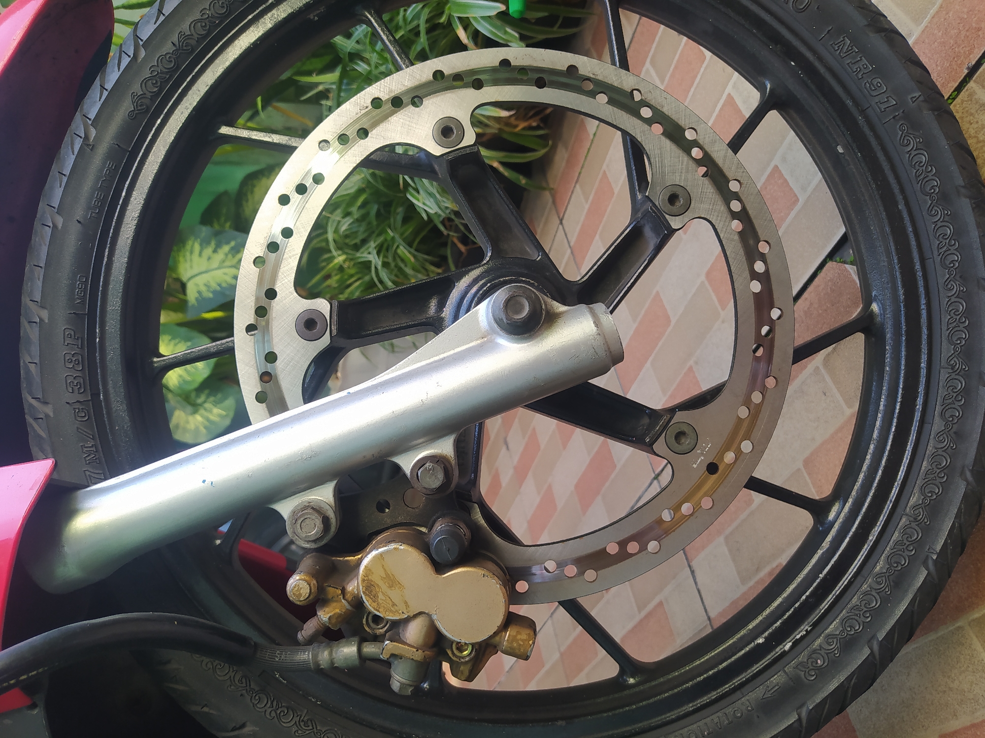 A close-up, eye-level shot captures the front wheel, brake disc, and part of the suspension of a red motorcycle. The motorcycle is parked on a tiled floor with a pattern of beige and red squares. In the background, lush green foliage is visible, suggesting an outdoor setting. The image appears to be taken in Sroyo, Indonesia, given the commonality of red motorcycles and tiled outdoor areas in residential or communal spaces there. No people are present in the image.