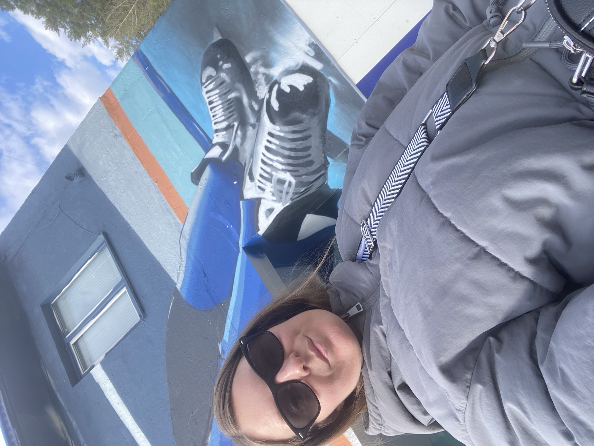 This outdoor photograph is taken at a tilted angle, capturing a person against a vibrant mural on a building wall under a bright sky. In the foreground, a woman with long brown hair is visible, wearing black cat-eye sunglasses and a grey, puffy winter jacket. A black handbag with a distinctive black and white patterned strap is seen over her shoulder. She appears to be looking towards the camera with a neutral expression.
The background features a building facade painted in shades of blue and grey, adorned with a large mural. The mural prominently displays two grayscale, vintage-style microphones with stylized white highlights, set against a backdrop of light blue, orange, and darker blue stripes. To the left of the mural, a rectangular window with venetian blinds is set into the wall. Above the building, a clear blue sky with scattered white clouds and the tops of green trees are visible, indicating a daytime setting. The image portrays a casual outdoor moment in what is identified as Noviy, Russia. There is no visible text in the image.