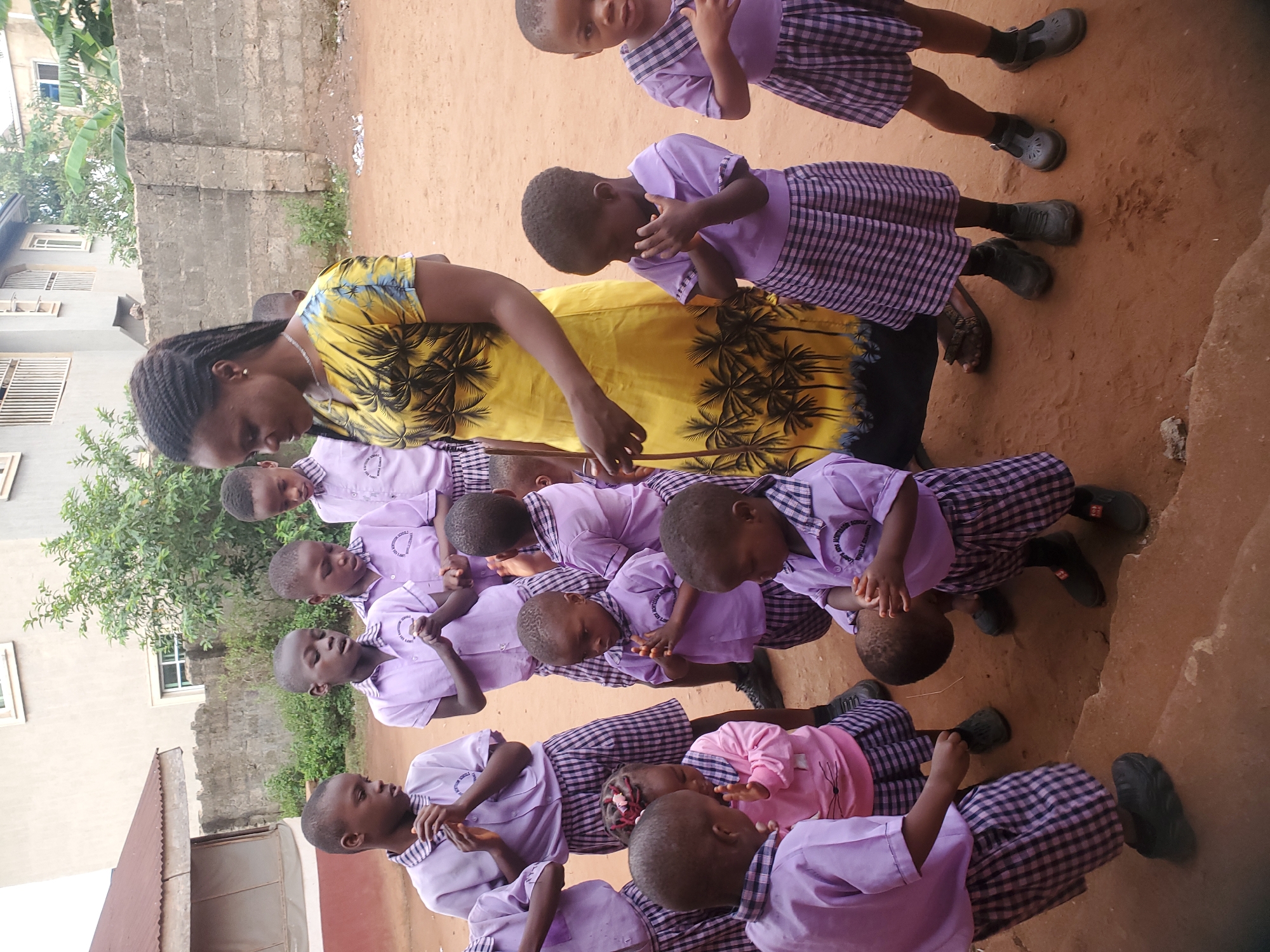 In Ukwu-Abwa, Nigeria, a group of young children, likely primary school students, are gathered outside on a dirt ground, possibly at their school. They are dressed in a uniform consisting of purple shirts and plaid skirts or shorts. A woman wearing a yellow dress with a palm tree print is standing among the children, possibly instructing or assisting them with an activity involving their hands, which they have raised near their faces. A building is visible in the background.