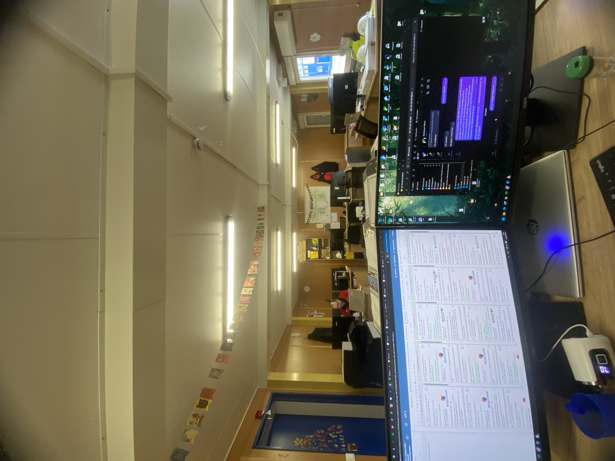 The image shows an office environment with multiple computer monitors displaying various programs and applications. The perspective is slightly tilted and the scene extends into the background, showing more desks and office equipment.
The foreground is dominated by computer monitors, a laptop, and office supplies such as a mouse and a small electronic device showing a digital reading of "70." The screens display what seems to be work-related applications. One shows a messaging application with purple text bubbles, and the other displays tables filled with data and text in Russian.
In the background, there's a glimpse into other parts of the office. People are working at desks, and there's a banner in the center of the image. Decorations hang from the ceiling.
There is no definitive information to confirm the location in the city of Luzhitsy, Russia.
