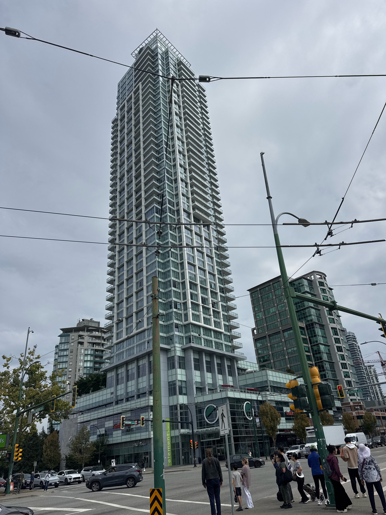 This is a full-shot, eye-level, outdoor, daytime, medium-low angle photograph of a busy intersection in Burnaby, Canada. The scene is dominated by a very tall, modern, multi-story residential building with a white exterior and numerous balconies. In the foreground, a green street pole supports traffic lights, power lines, and streetcar wires. Several cars, including an SUV, sedan, and smaller vehicles, are visible on the road. Pedestrians, some with shopping bags or pushing strollers, are waiting at the crosswalk or walking on the sidewalk. Other modern buildings, some with glass facades, are visible in the background, along with trees and utility poles. The sky is overcast and grey. The overall atmosphere is that of an urban environment.