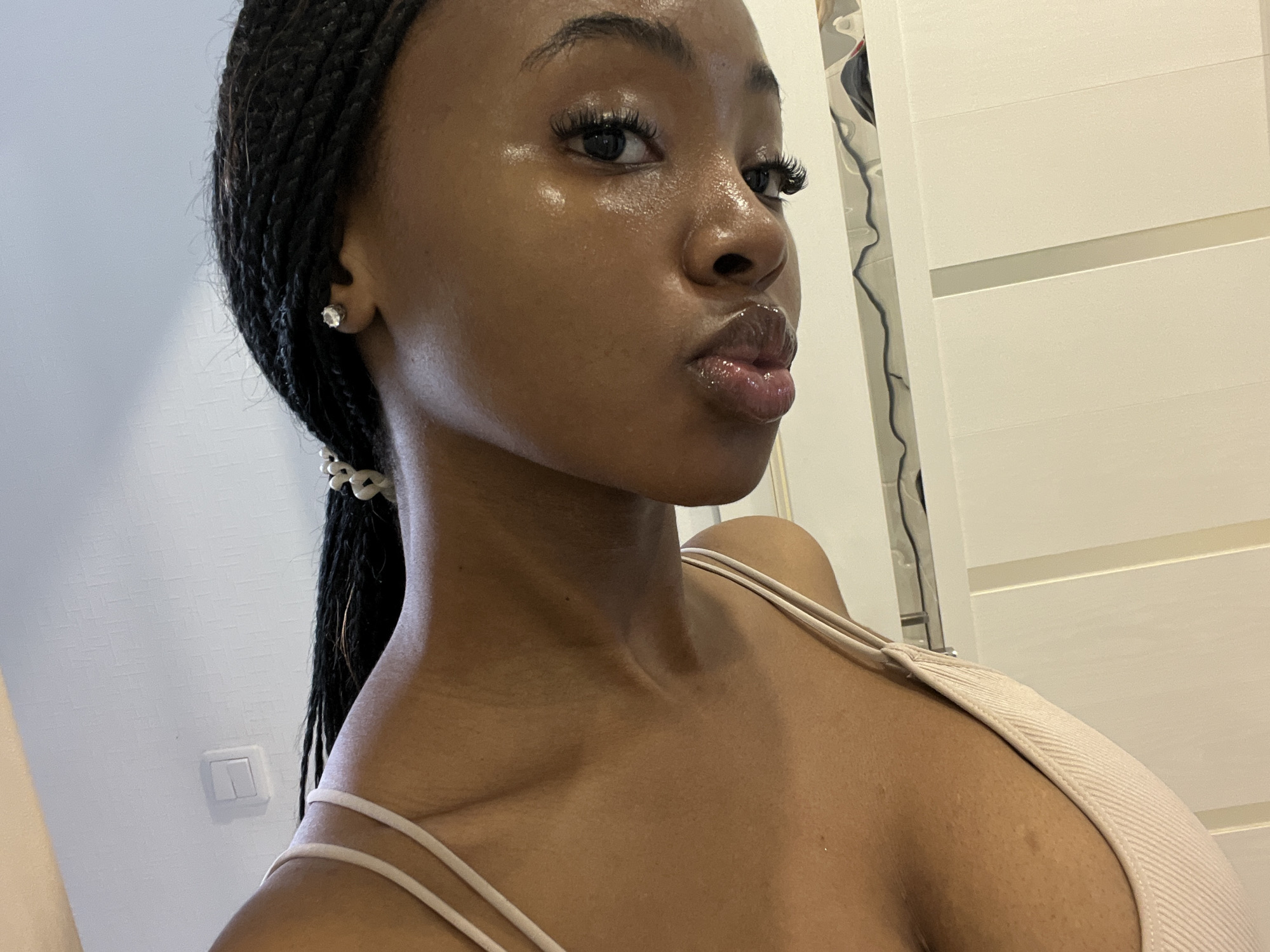 A close-up, head-and-shoulders photograph shows a young Black woman from the chest up, posing for a selfie. She is wearing a light-colored, thin-strapped top. Her dark hair is braided into a style that extends down her back. She has on stud earrings and her makeup includes lip gloss and mascara. The background is a white wall with a light fixture and a door with horizontal panels. The image is taken indoors, likely in a residential setting. There is no information in the image that would suggest a location in Kazan, Russia.