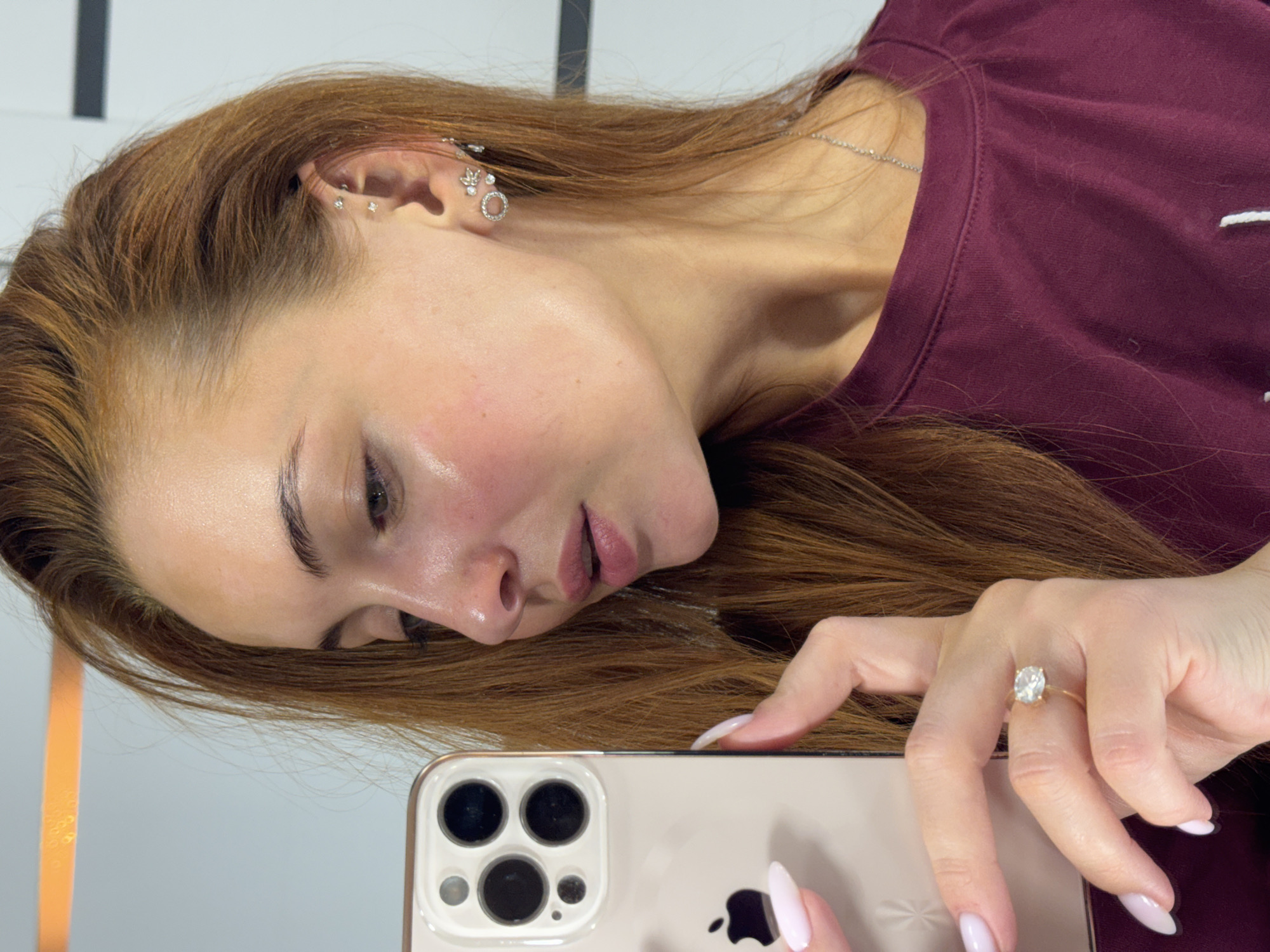 A woman is taking a selfie. She is holding a gold iPhone. She is wearing a maroon t-shirt and a gold ring with a large oval stone. She has several earrings in her left ear. The location appears to be indoors with a white wall with black and gold accents. I cannot verify if the location is in Chaikovsky, Russia.