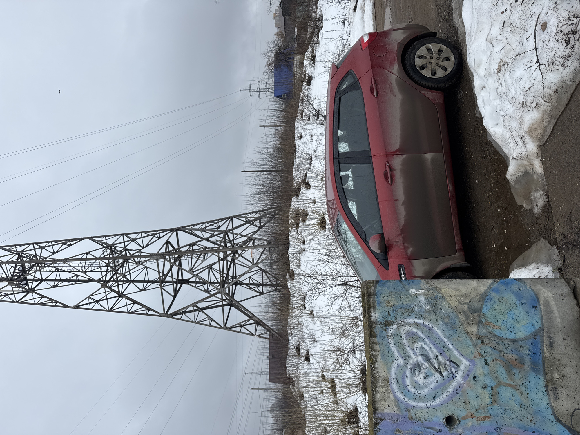 The image depicts a red hatchback car parked next to a concrete barrier with graffiti on a gray, overcast day. The vehicle is noticeably dirty, suggesting recent travel on unpaved roads. Patches of snow are visible in the background, indicating a cold climate. A large electrical transmission tower dominates the left side of the frame, with power lines stretching across the sky. The scene appears to be on the periphery of an urban area, with bare trees and vegetation interspersed with urban infrastructure. The location is likely near power lines and a road on the outskirts of Chaikovsky, Russia.
