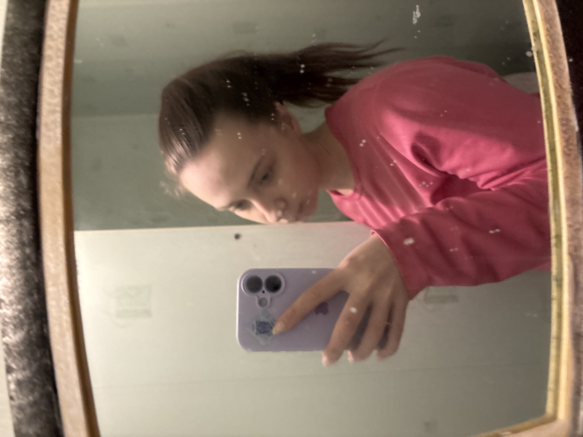 A young woman in a pink long-sleeved shirt takes a selfie in a mirror with a purple phone that has a flower sticker on its lower-left corner. The woman is looking down at her phone. The background appears to be a bathroom. The city of Прикамский, Russia is not directly discernible from the given image.