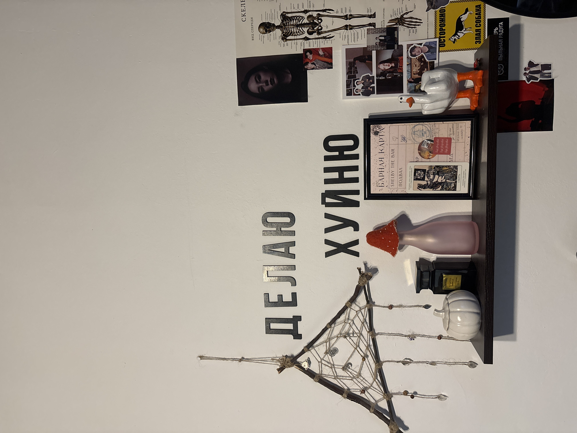 The image depicts a cluttered wall decorated with posters, a shelf, and decorative items. The scene likely takes place indoors, within a home or apartment. The wall is painted a light color. On the wall are two posters, one featuring a human skeleton and another with various images. A shelf is mounted on the wall, holding a framed piece of artwork that says "Shelby the Bar." It also holds a goose figurine giving the middle finger, and several other decorative objects, including a vase, a bottle of perfume, and a small pumpkin. Below the shelf hangs a dream catcher. Black block letters are placed on the wall spelling out Russian words vertically.
The location in Верхнеказымский, Russia is inferred from the Russian text on the wall.