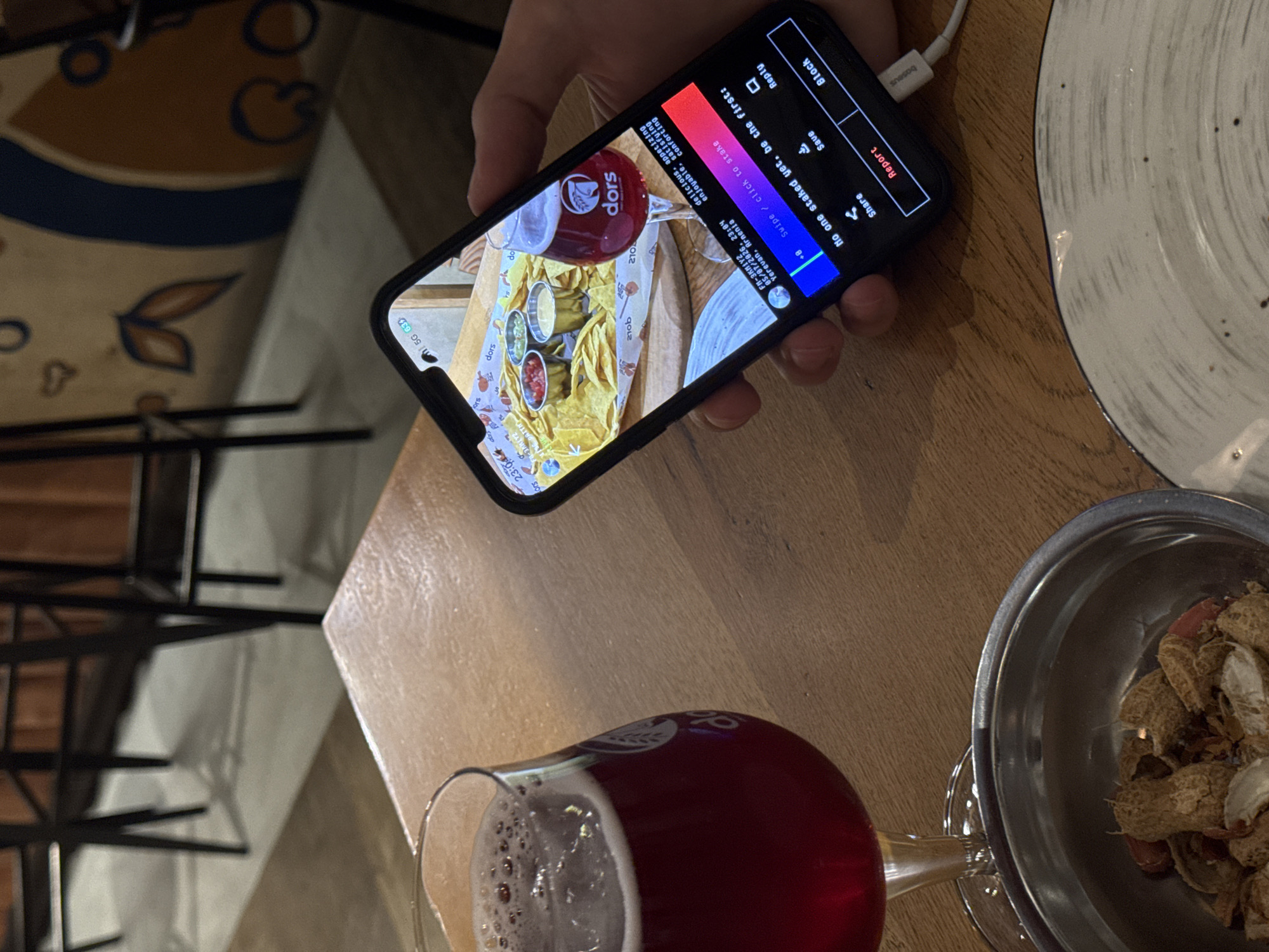A mobile phone held in a hand displays an image of a table setting featuring food and drink. The image displayed on the phone is a close-up of a tray of tortilla chips with multiple dipping sauces, alongside a red-colored drink in a glass. The drink is labelled with "Dors." The user interface of the mobile phone is in view, showing options to "Block," "Save," "Reply," and "Report."
The mobile phone is positioned above a real-life setting of a wooden table with a glass of red-colored liquid, possibly a beverage, and a silver bowl containing peanut shells. Next to these is the corner of a ceramic plate with speckles.
The OCR data suggests a date and time (05/07/2026, 23:04) and a location: Yerevan, Armenia. This could be indicative of the location where the image was captured or where the displayed image on the phone was originally taken.
