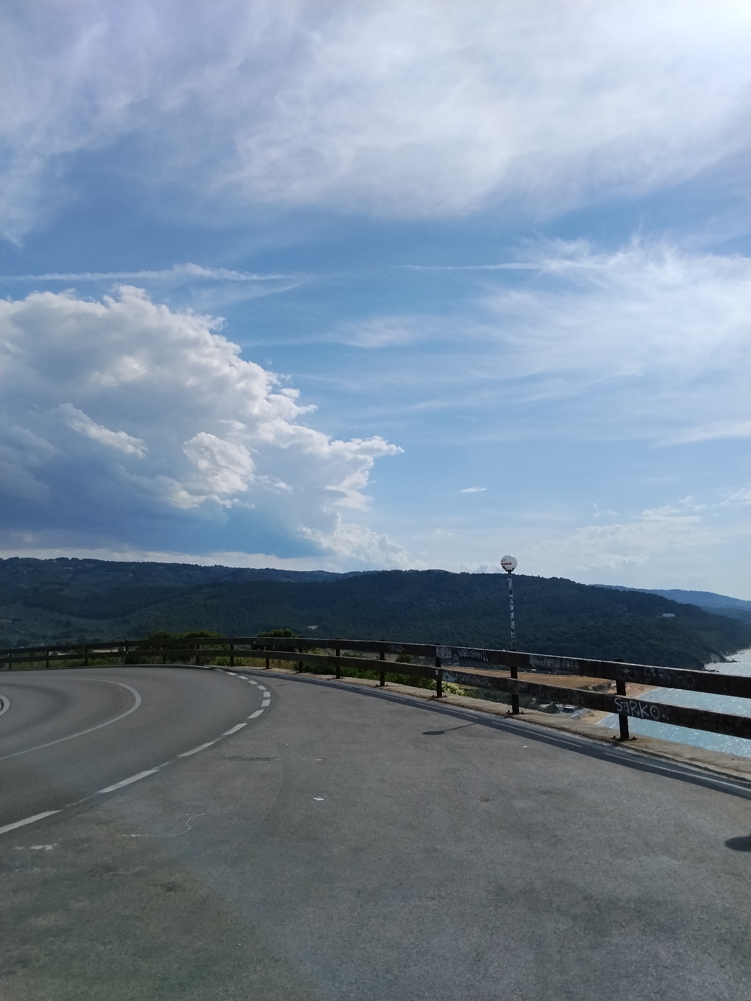 The image presents a panoramic view from a paved coastal road under a partly cloudy sky, characteristic of Vico del Gargano, Italy. In the foreground, an asphalt road curves to the left, marked by a solid white line on its inner edge and a dashed white line along its right edge. Parallel to the road on the right is a multi-bar wooden railing, heavily marked with graffiti including the legible word "SARKO." Beyond this railing, a densely forested hillside covered in green trees slopes downwards. A tall, slender pole topped with a white spherical object stands vertically on this hillside, approximately halfway up. To the far right, the blue-green waters of the Adriatic Sea are visible, with a narrow stretch of beach at the base of the hill, where several small, light-colored structures (possibly beach facilities) are discernible. In the mid-distance on the left, more forested hills extend. The sky occupies the upper half of the image, displaying a gradient of blue with large, white cumulus clouds concentrated on the left and more wispy, high-altitude clouds with faint contrails scattered across the right. No individuals, vehicles, or explicit human activities are present in the scene.