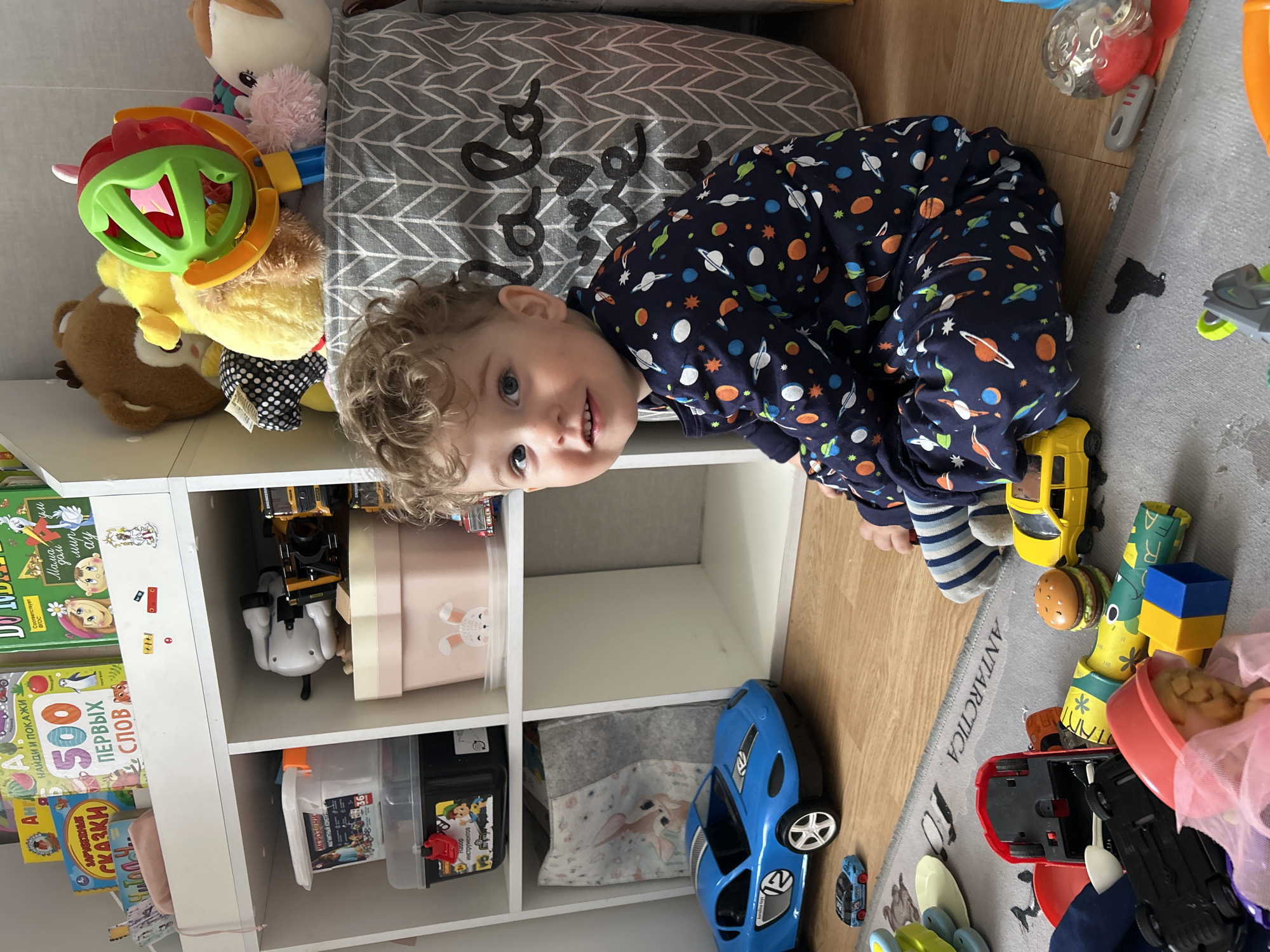 A child wearing a navy blue pajama set with planet and star designs is kneeling on a gray rug with the word "ANTARCTICA" printed on it. The child is looking at the camera with a slight smile. The rug is positioned on a light wood floor. Behind the child is a white shelving unit filled with various toys, books, and storage boxes. Toys include stuffed animals, toy cars, and building blocks. The books appear to be children's books, including one with the title "500 ПЕРВЫХ СЛОВ" (500 First Words). The location is Klin, Russia.