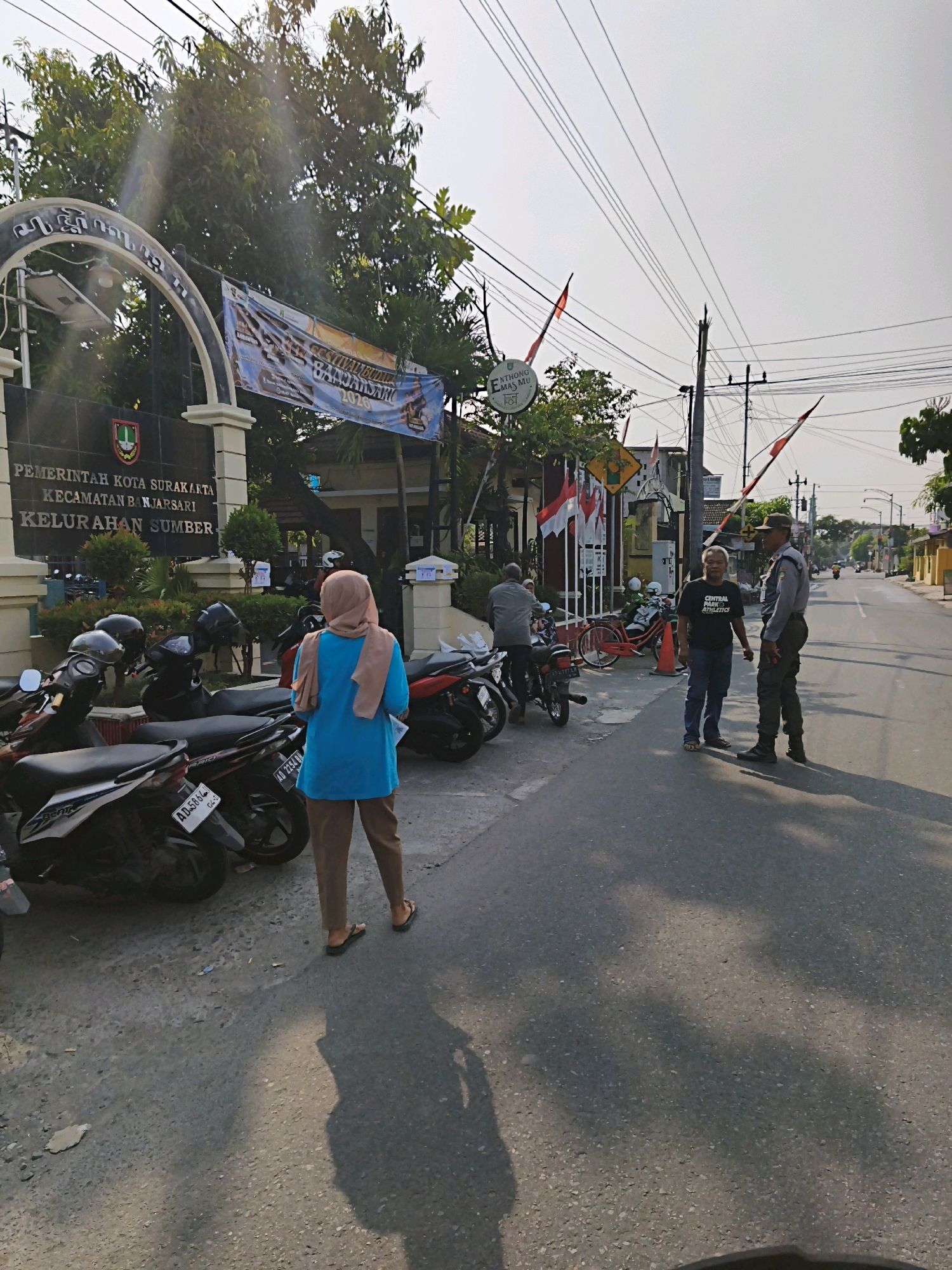 The image depicts the exterior of a government building in Surakarta, Indonesia, likely a sub-district office or similar administrative facility, as indicated by the signage reading "PEMERINTAH KOTA SURAKARTA KECAMATAN BANJARSARI KELURAHAN SUMBER" (Government of Surakarta City, Banjarsari District, Sumber Village). The building has a dark facade with a large arched entranceway. A banner with Indonesian flag colors and text about "FESTIVAL BUDAYA BANJARSARI 2020" hangs from the entrance.
In the foreground, a collection of motorcycles is parked on the side of a paved road. A woman wearing a light blue long-sleeved shirt, a beige hijab, and dark pants, stands with her back to the camera, facing the building, holding some papers. Further down the road, a man in a black t-shirt and blue jeans, and another man in a police-like uniform with a hat and dark pants, are standing and conversing. The environment suggests an outdoor, daytime setting with clear skies and a few trees visible. Electrical wires are prominent in the upper portion of the image, crossing the sky. The overall scene conveys a snapshot of daily activity outside a local government office.