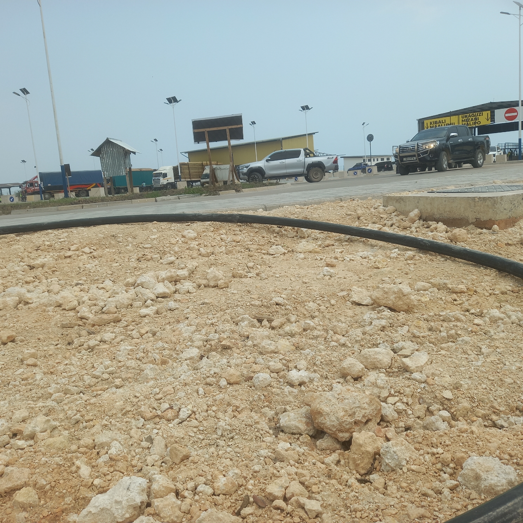 In Bububu, Tanzania, a construction site is visible with various vehicles and infrastructure. In the foreground, a pile of sandy dirt and rocks, with a black pipe curving across the scene. In the mid-ground, a silver pickup truck is parked. Behind it, a series of buildings and structures are present, including a yellow warehouse-like building and a smaller, rustic shelter. Several large trucks, including a white semi-truck and a blue truck with a trailer, are also visible. In the background, streetlights with solar panels dot the landscape, and a sign with writing in a foreign language is mounted on a structure. The sky is overcast.
