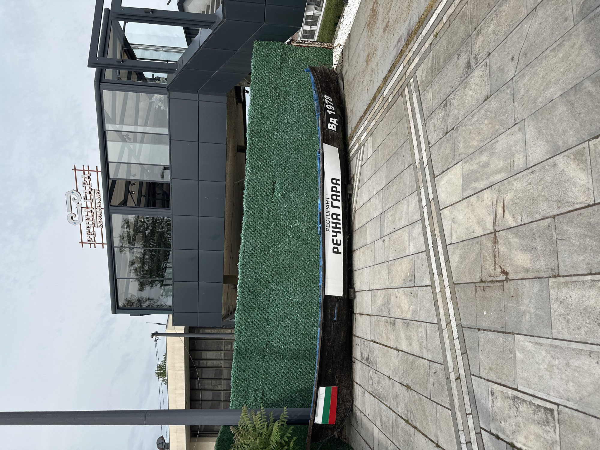 A boat with the name "Речната Гара" and the year "Вд 1978" on its side rests on a paved area. A green decorative hedge partially fills the boat's interior. A Bulgarian flag is affixed near the bow. The boat is situated next to a modern building with large windows and a grey facade. The surrounding pavement is composed of rectangular stone slabs. A streetlight is also visible in the frame. This photograph shows a decorative boat at the River Station in Vidin, Bulgaria.