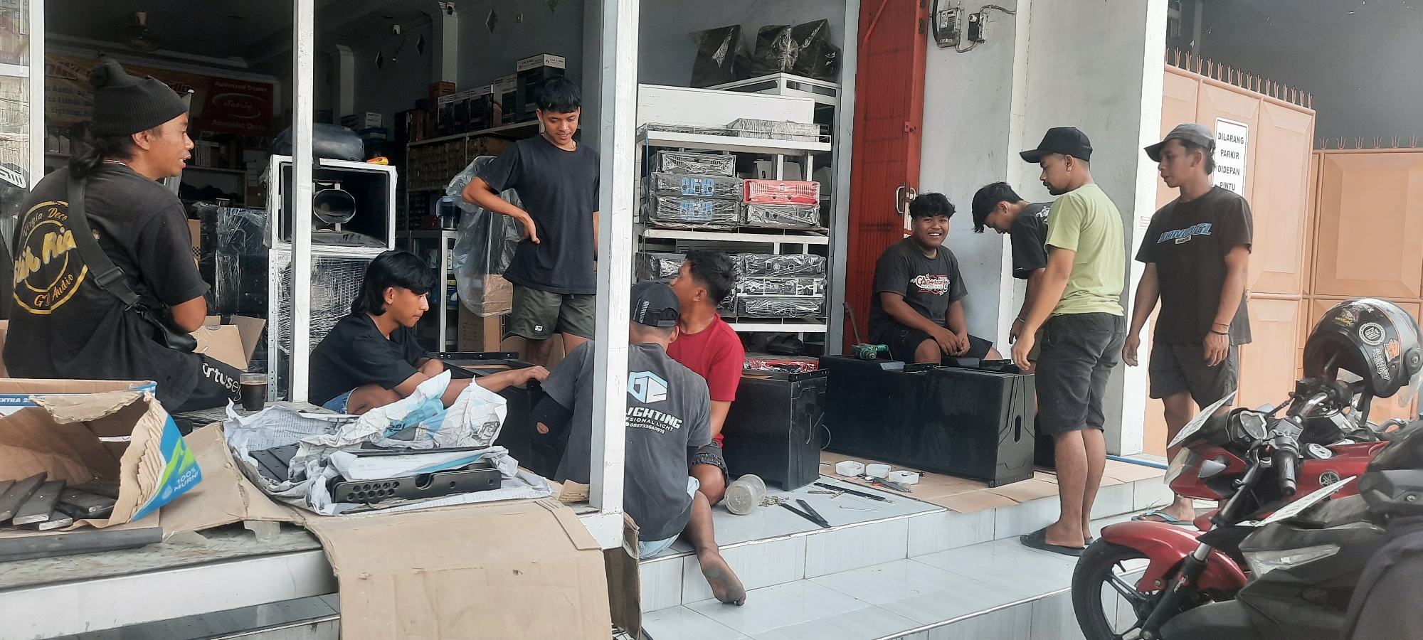 In front of an outdoor shop in Selopuro, Indonesia, a group of eight young men are gathered, some appearing to be working with audio equipment. The shop's interior displays rows of audio processors and other electronic equipment. In the foreground, several large black speaker cabinets are arranged on the ground, and one is open, revealing its internal components. A man wearing a black beanie and a black t-shirt with yellow lettering sits near the front, holding a drink. Another young man with a mullet hairstyle sits on the ground next to a speaker. Other men are standing or sitting around the speaker cabinets, engaged in conversation or looking at the equipment. A red motorcycle with a helmet is parked to the right of the scene. The overall impression is of a casual work environment for a group of young men dealing with sound systems.
