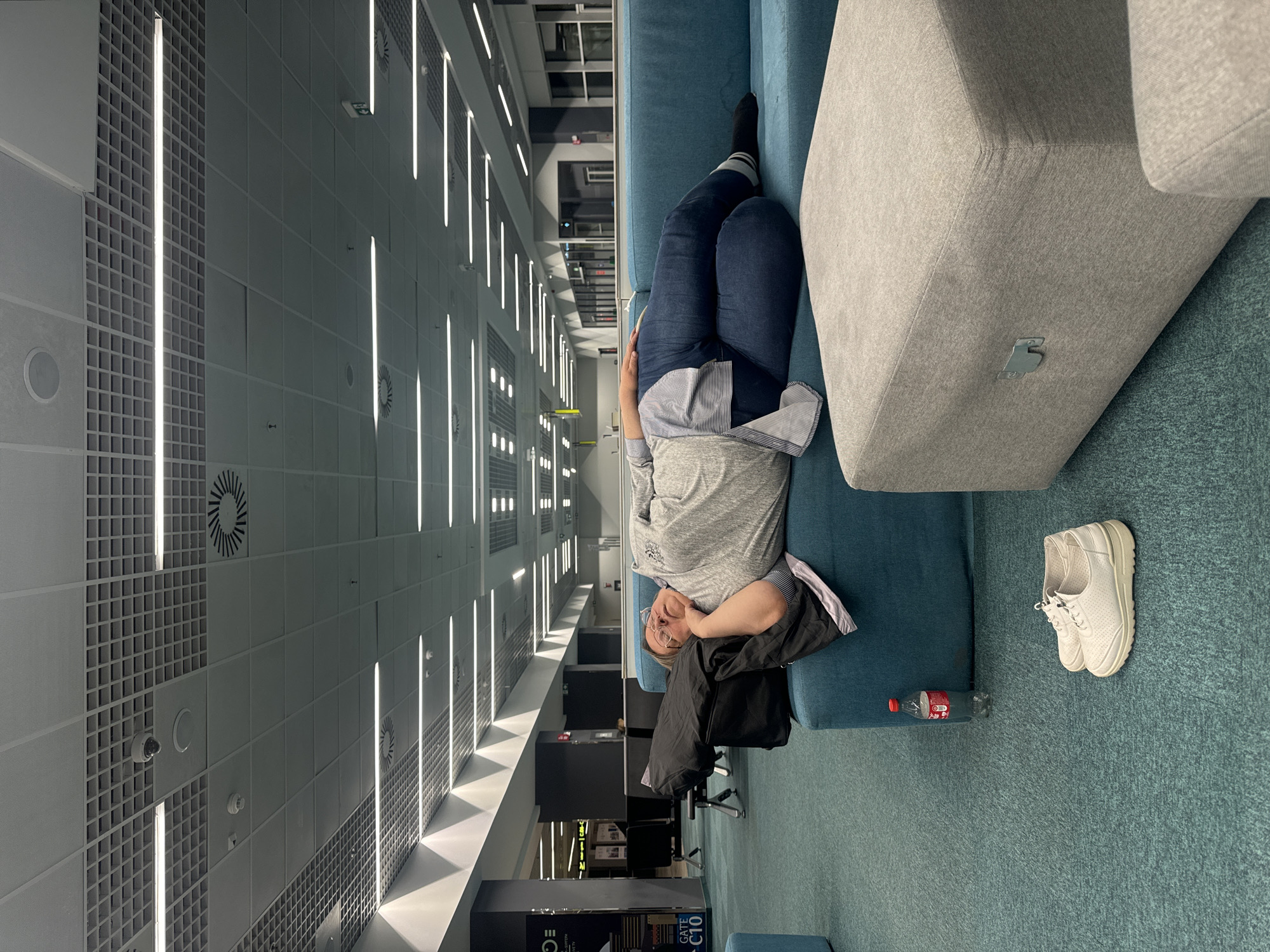 The image shows a person lying on a blue sofa in what appears to be a large, modern building. The person is wearing blue jeans, a gray shirt, and a striped shirt underneath. They have glasses on and are facing the camera. A black jacket is draped near the person, and a bottle of soda and a pair of white sneakers are on the floor beside the sofa. The building features a grid-patterned ceiling with long, linear light fixtures. In the background, there are computer stations, bookshelves, and windows indicating a multi-story structure. The location seems like a library or study area.